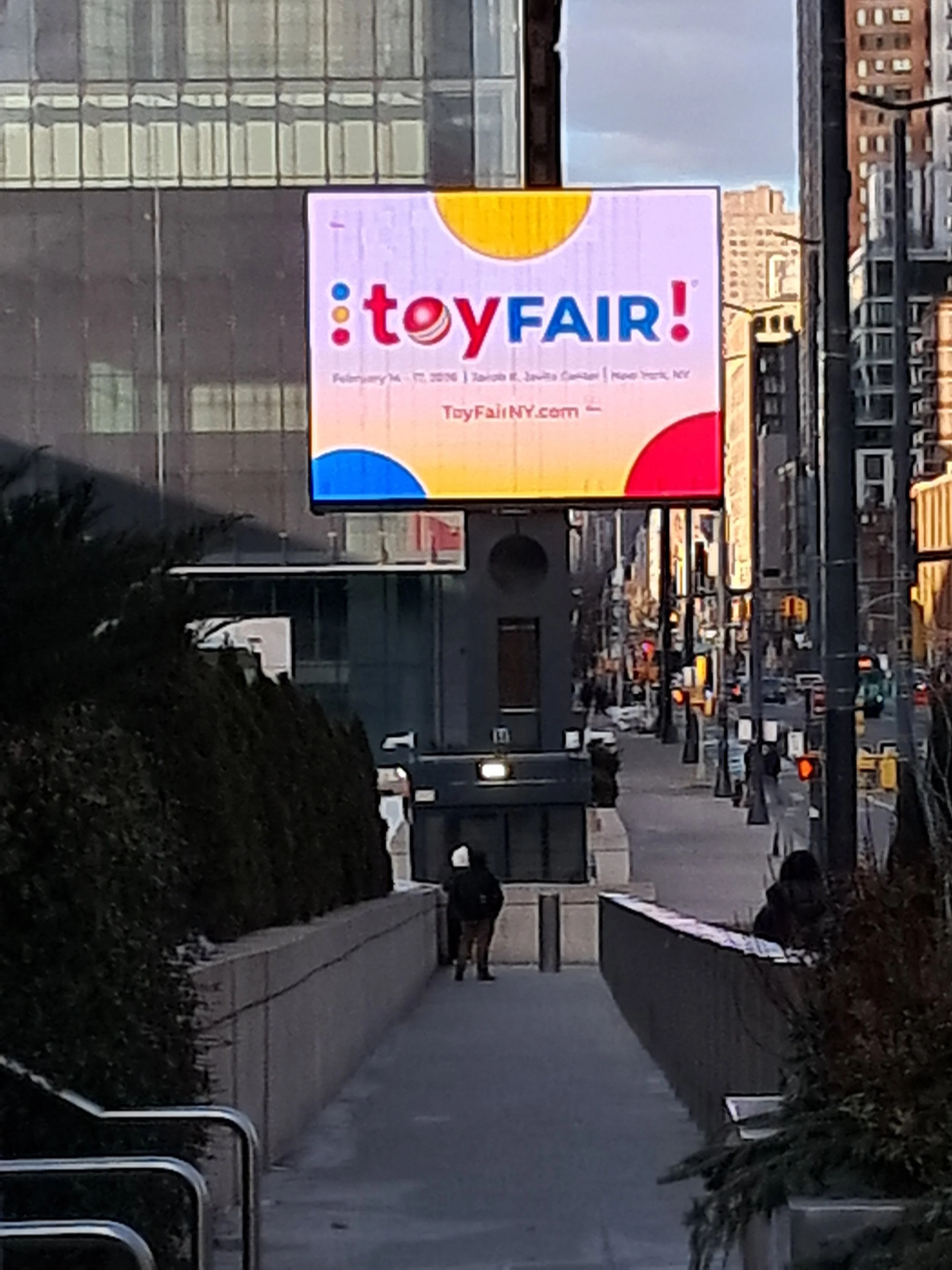 A city street with a billboard advertising Toy Fair, featuring colorful text and graphics, and surrounding tall buildings with glass windows. Pedestrians are walking along the sidewalk.