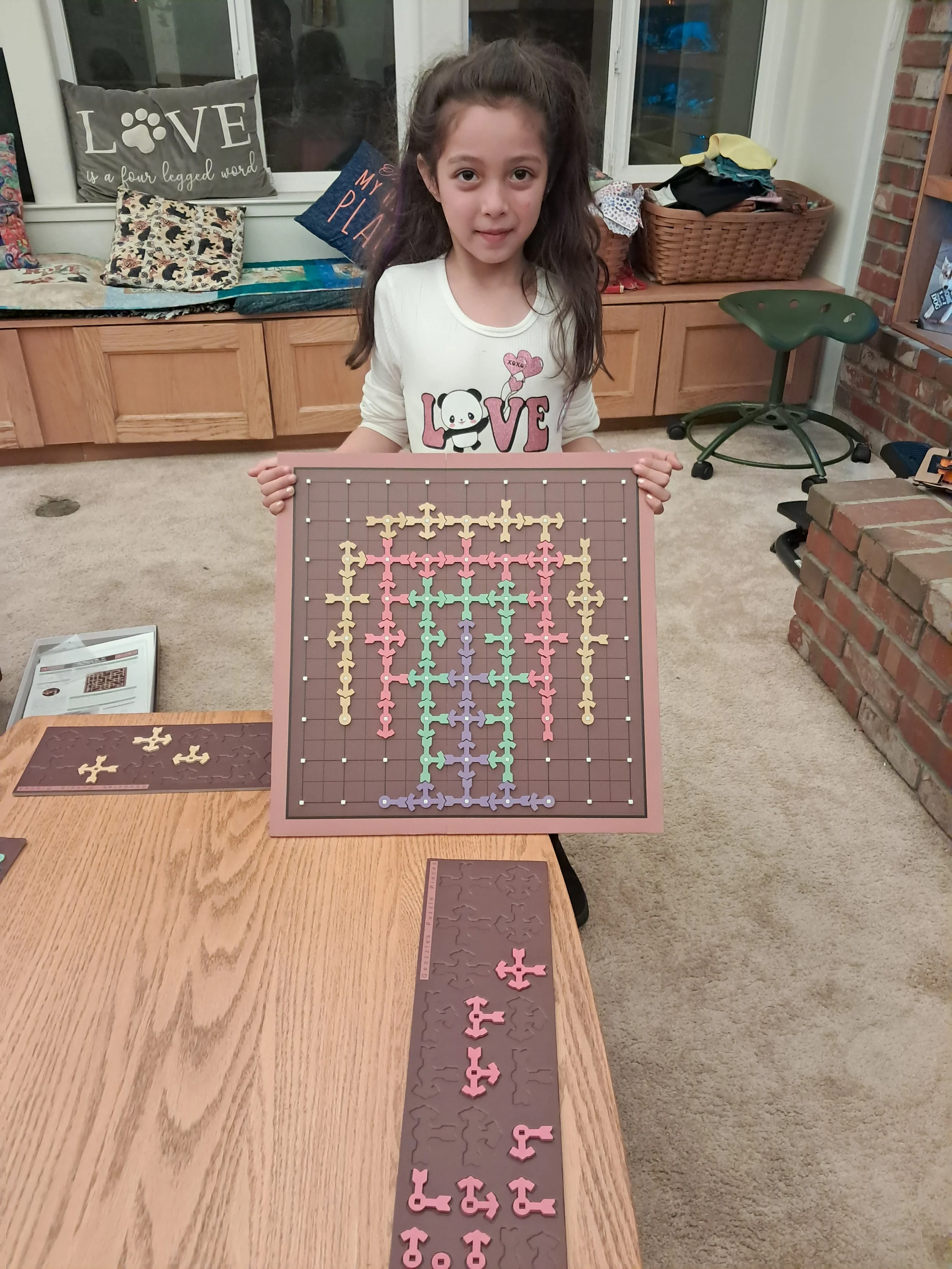 A young girl with brown hair and a white T-shirt featuring a panda and the word "LOVE" is holding a large, decorated game board with pink, yellow, green, purple, and beige pieces arranged on a gridded surface. She is standing behind a wooden table in a room with a brick wall and a window with a bench, pillows, and a wicker basket in the background.