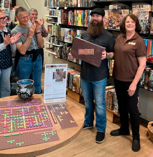 Group of people in a bookstore celebrating an event, with a man holding a board game titled Gozzle, a table with a colorful puzzle, and shelves of books in the background.