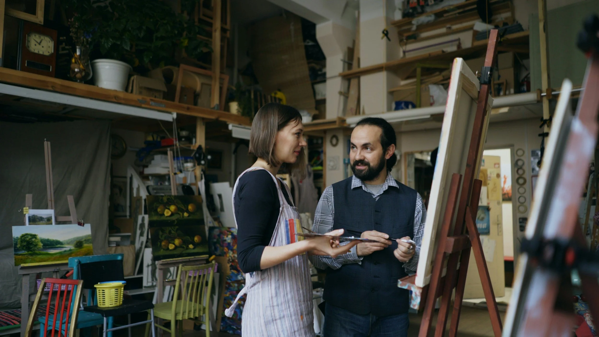 A woman and man speaking in an art studio surrounded by easels, paintings, and art supplies, with shelves and chairs in the background.