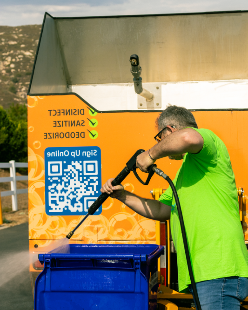 Man in a bright green T-shirt cleaning or disinfecting a public bike-sharing station or kiosk with a high-pressure water spray.