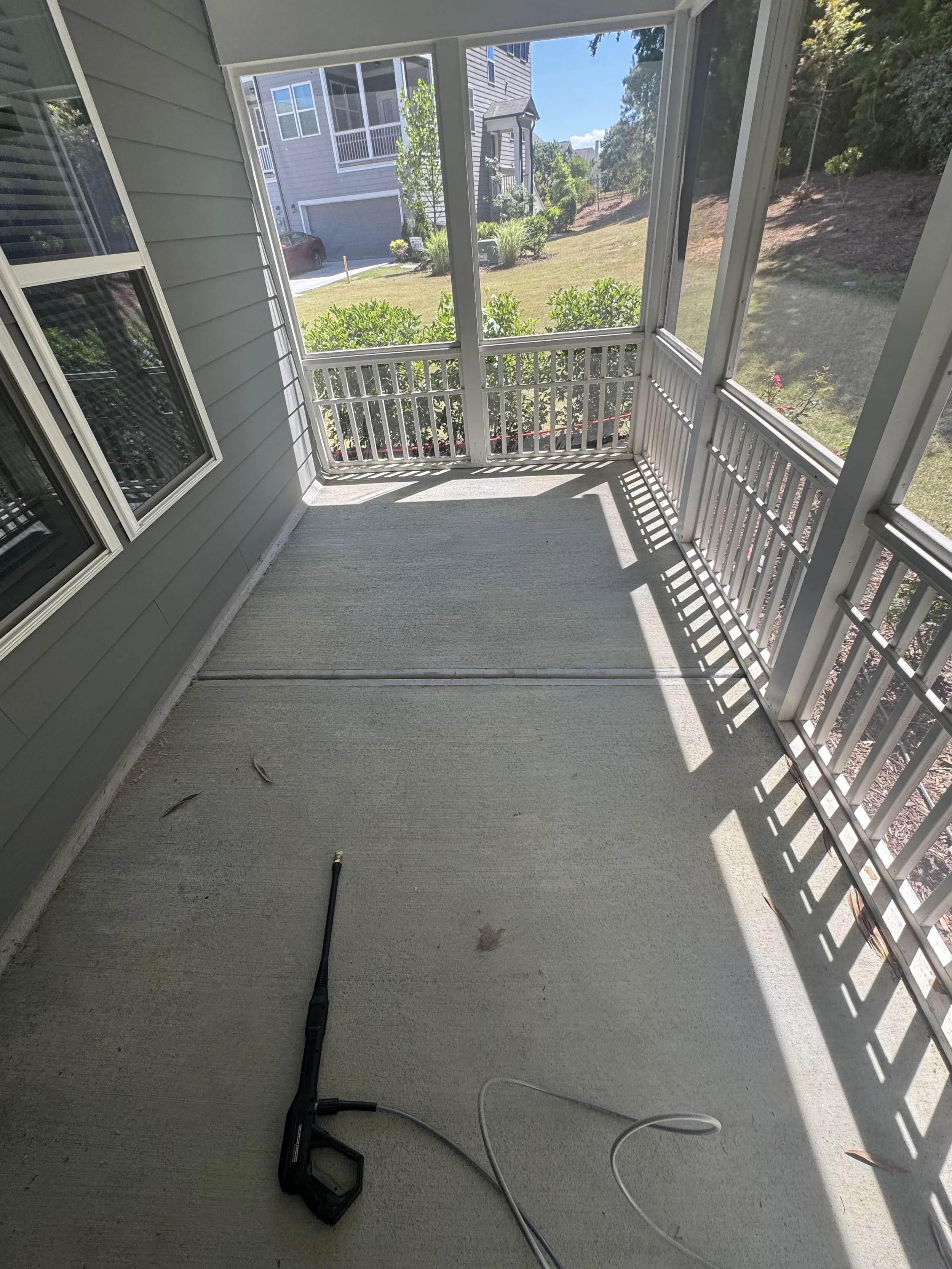 View from a porch or balcony showing a gray house with a garage, a red car, a lawn, shrubs, and neighboring houses in the background. Shadows cast by the railing create patterns on the floor.