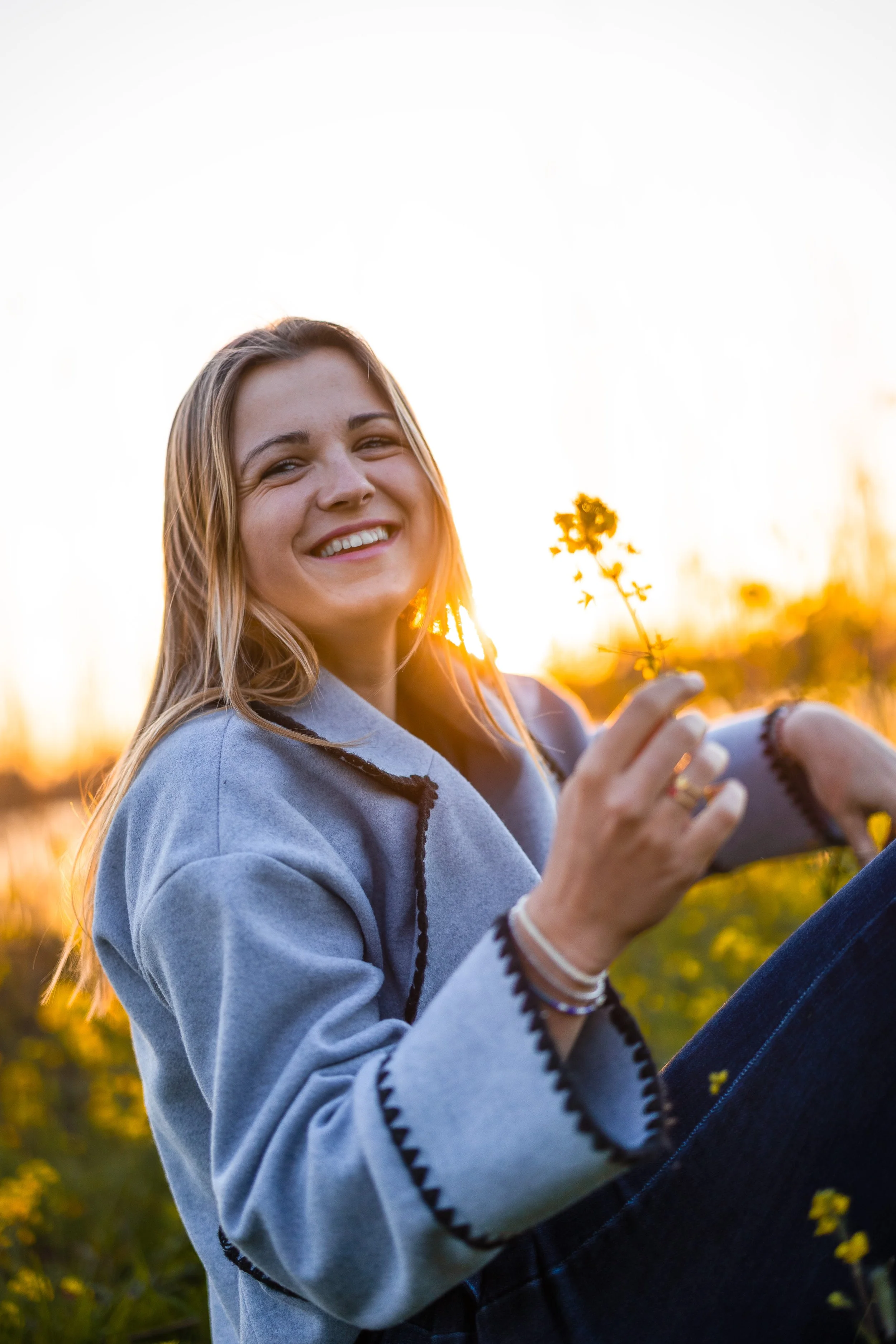 Vrouw die op de grond in een veld met gele bloemen zit en glimlacht, terwijl ze een bloem vasthoudt, bij zonsondergang.