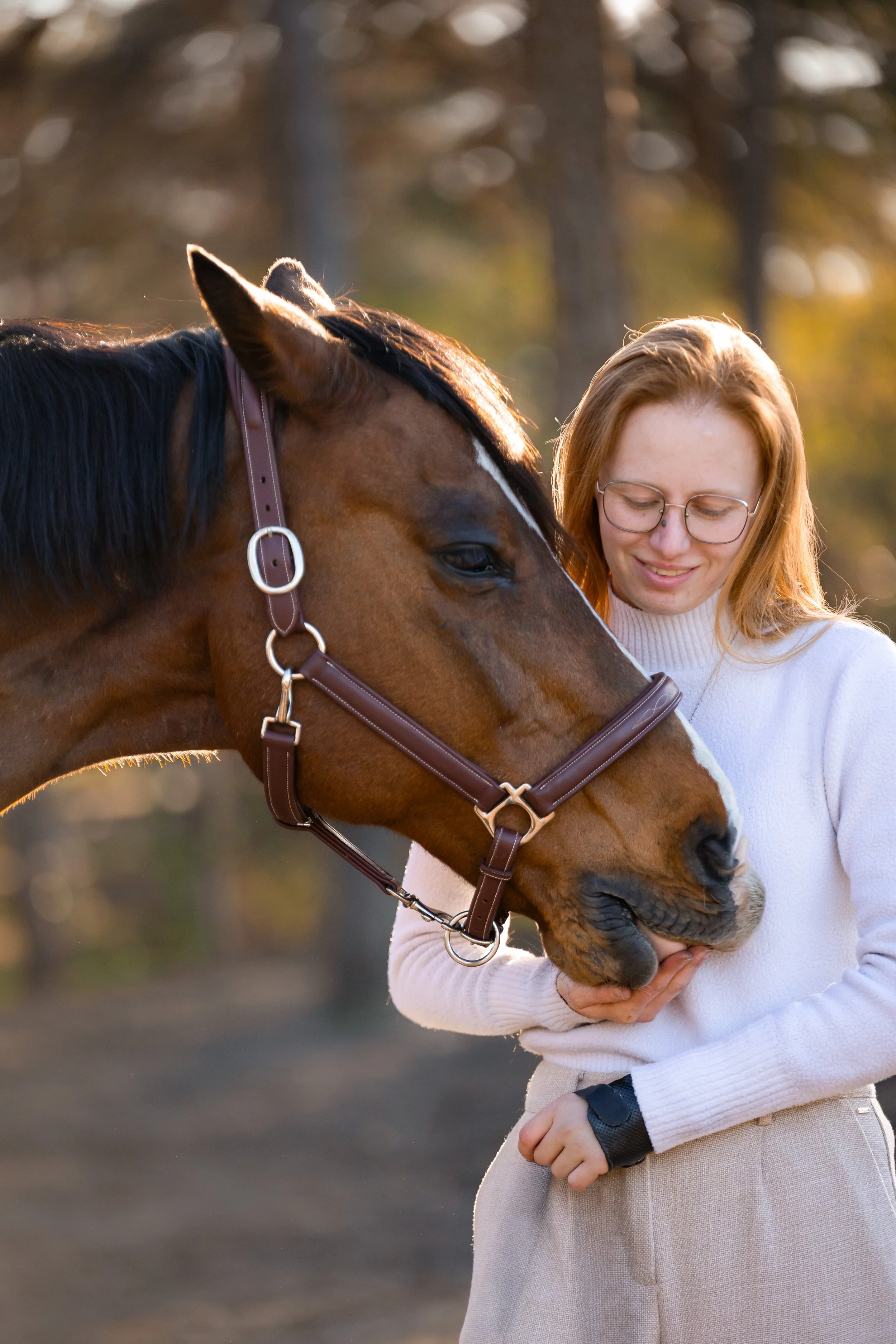 Een vrouw met rood haar en een witte trui staat met een paard in een bos, tijdens zonsondergang, en ze kijkt liefdevol naar het paard.