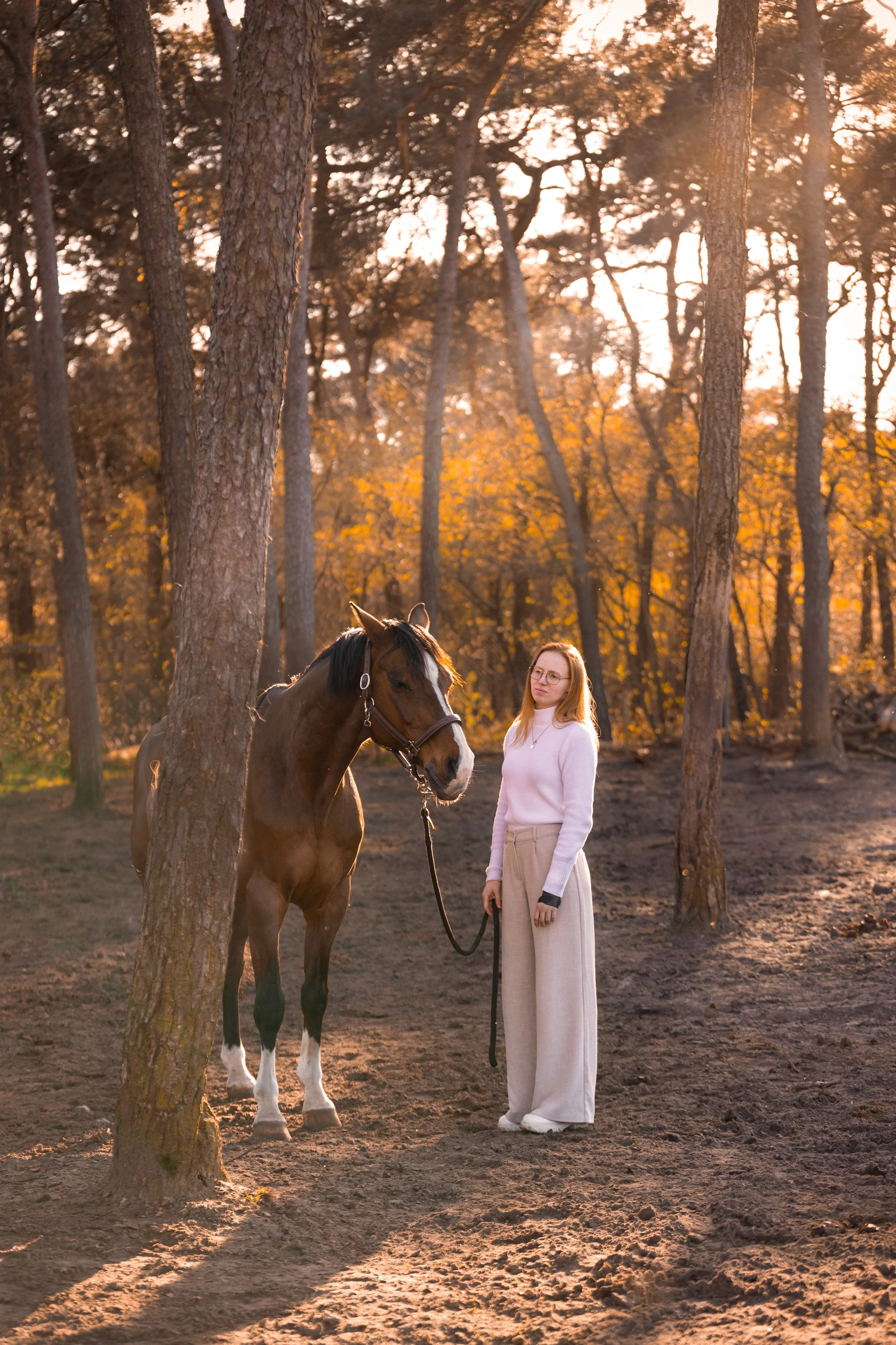 Een jonge vrouw in een wit trui en wijde broek staat naast een paard in een bos tijdens zonsondergang.