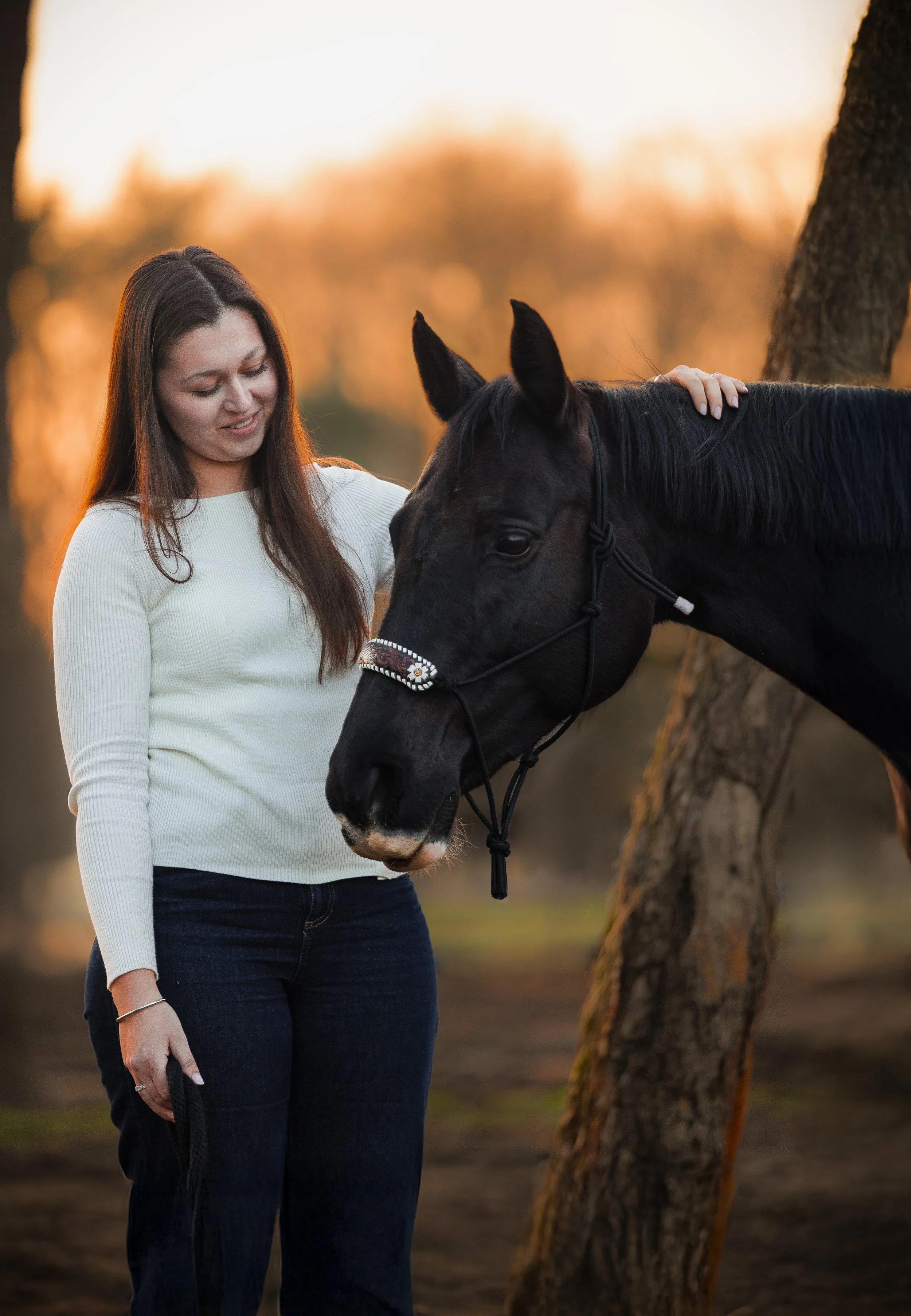 Een jonge vrouw met bruin haar in een witte trui staat naast een zwarte paard in een bos tijdens zonsondergang.
