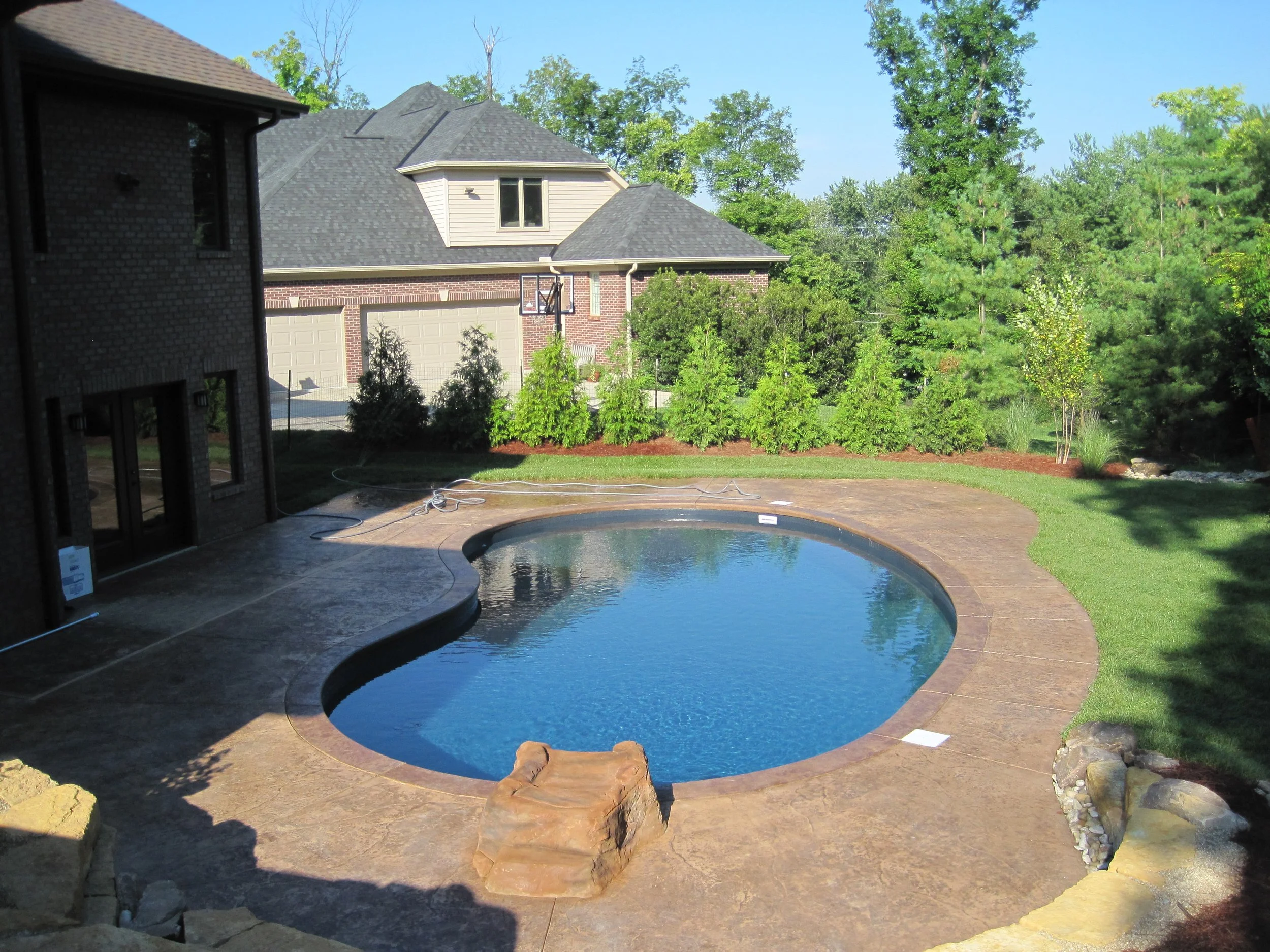 Backyard view with in-ground kidney-shaped swimming pool, surrounded by concrete patio and landscaped bushes near a residential house with a garage.
