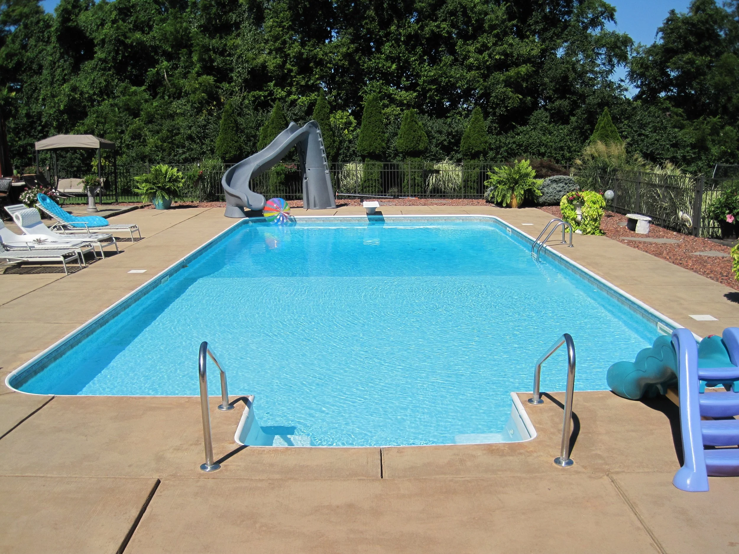 Outdoor swimming pool with diving board and slide, surrounded by lounge chairs and lush greenery.