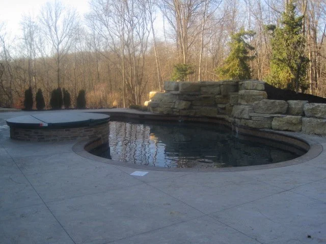 Backyard with a small, stone-surrounded swimming pool and spa. The pool area is bordered by a patio, and there are trees and rocks in the background.