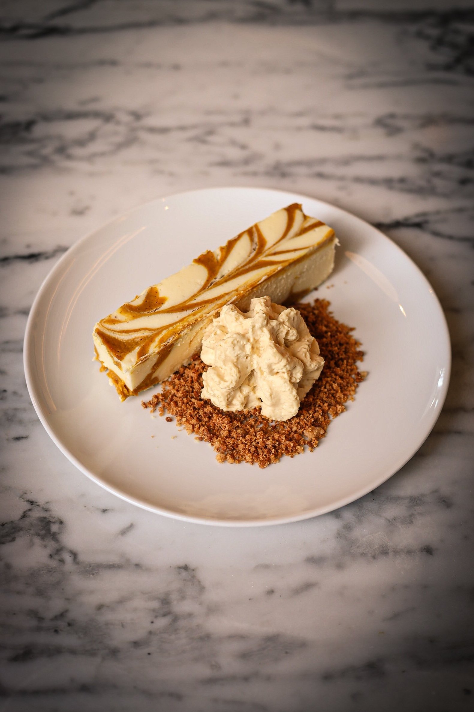 Slice of cheesecake with caramel and white chocolate swirl topping, served with whipped cream on a graham cracker crust, on a white plate.