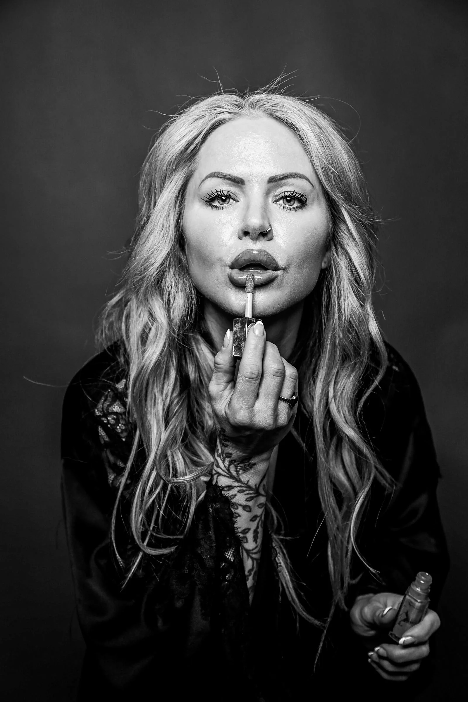 Black and white photo of a woman applying lipstick, holding a lipstick tube in one hand, with long wavy hair and tattoos on her arm, against a plain background.