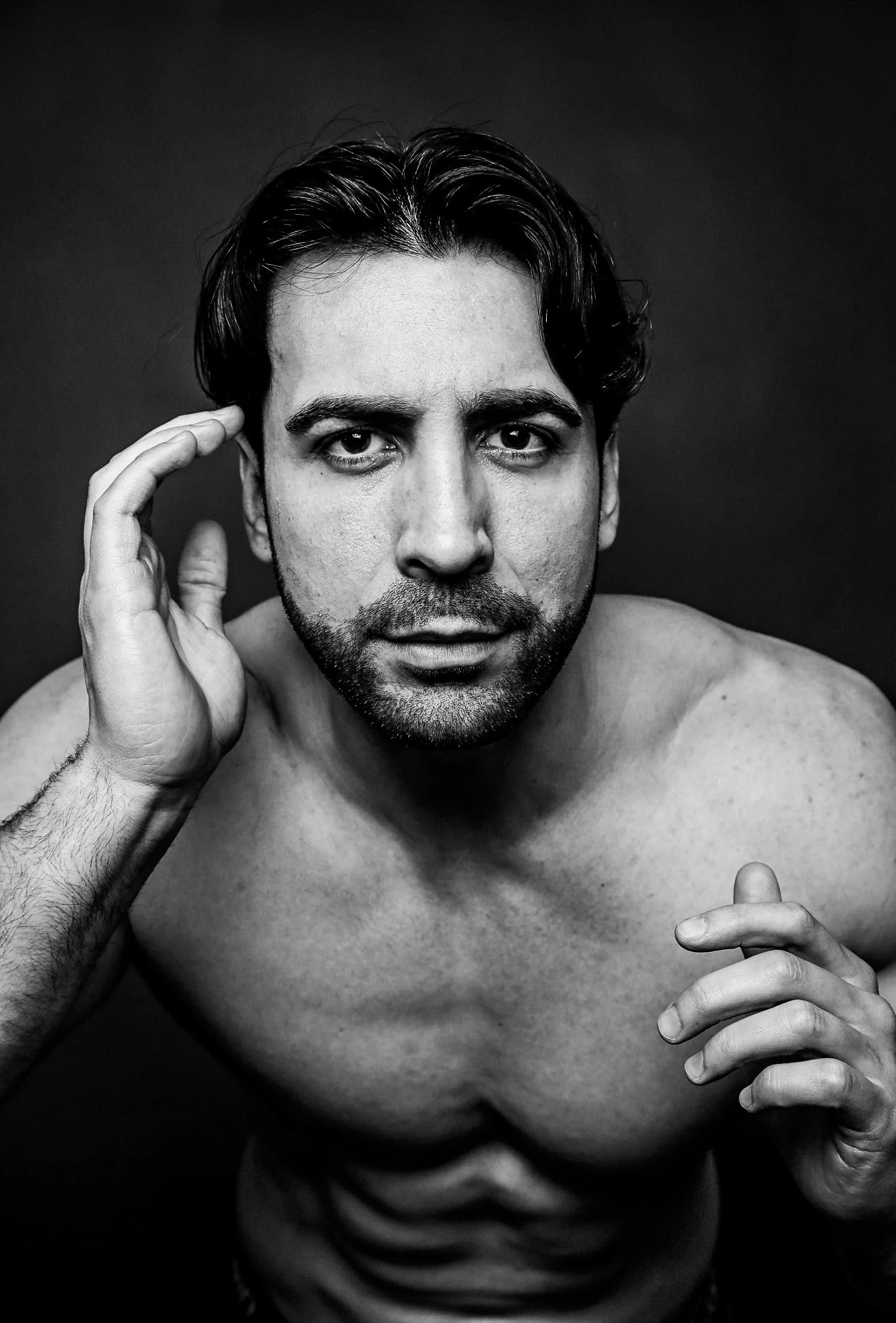 Black and white portrait of a shirtless man with medium-length wavy dark hair, looking directly at the camera, resting his right hand near his temple and slightly smiling.