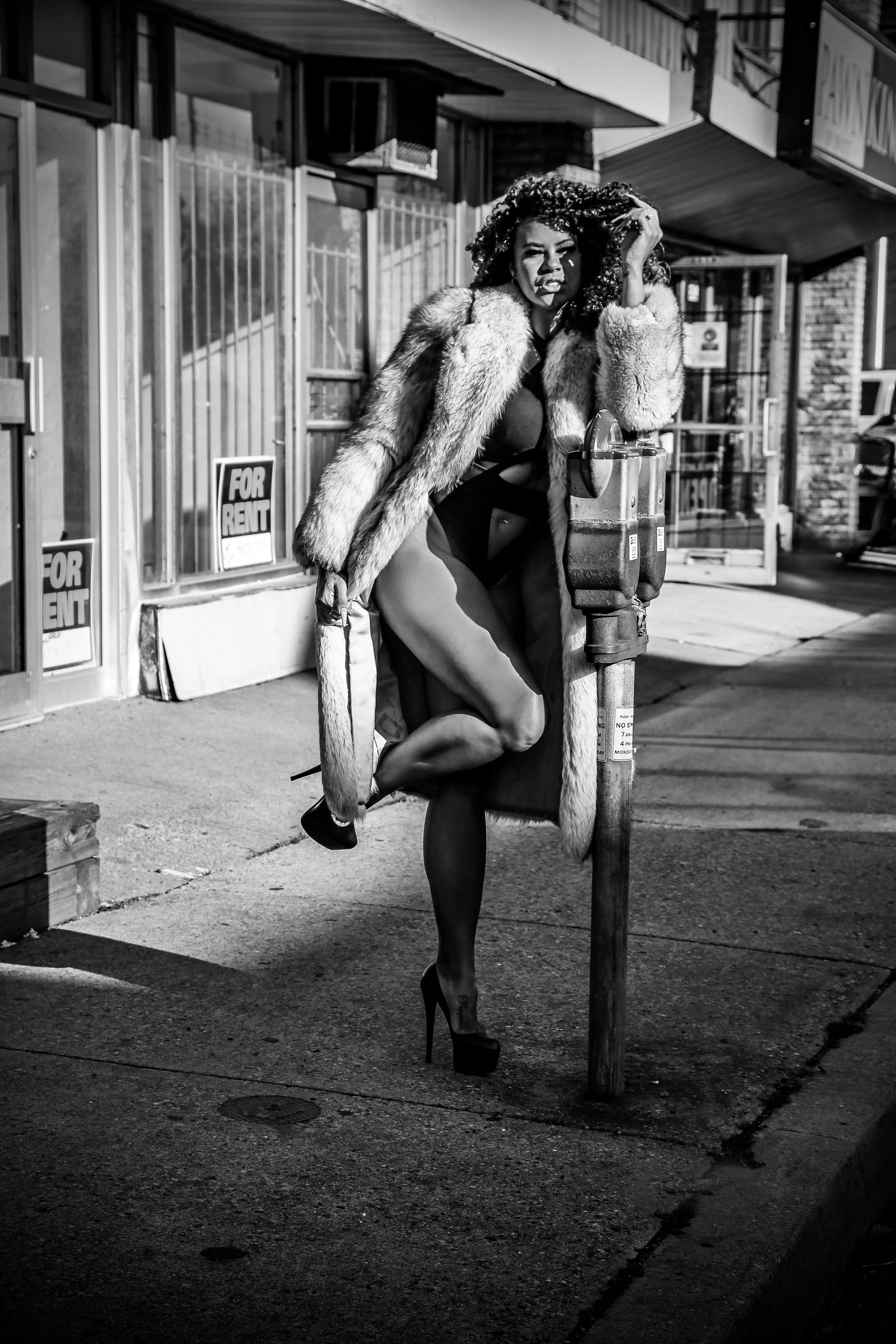 Black and white photo of a woman in high heels wearing a fur coat, leaning against a fire hydrant on a city sidewalk with storefronts and "For Rent" signs in the background.