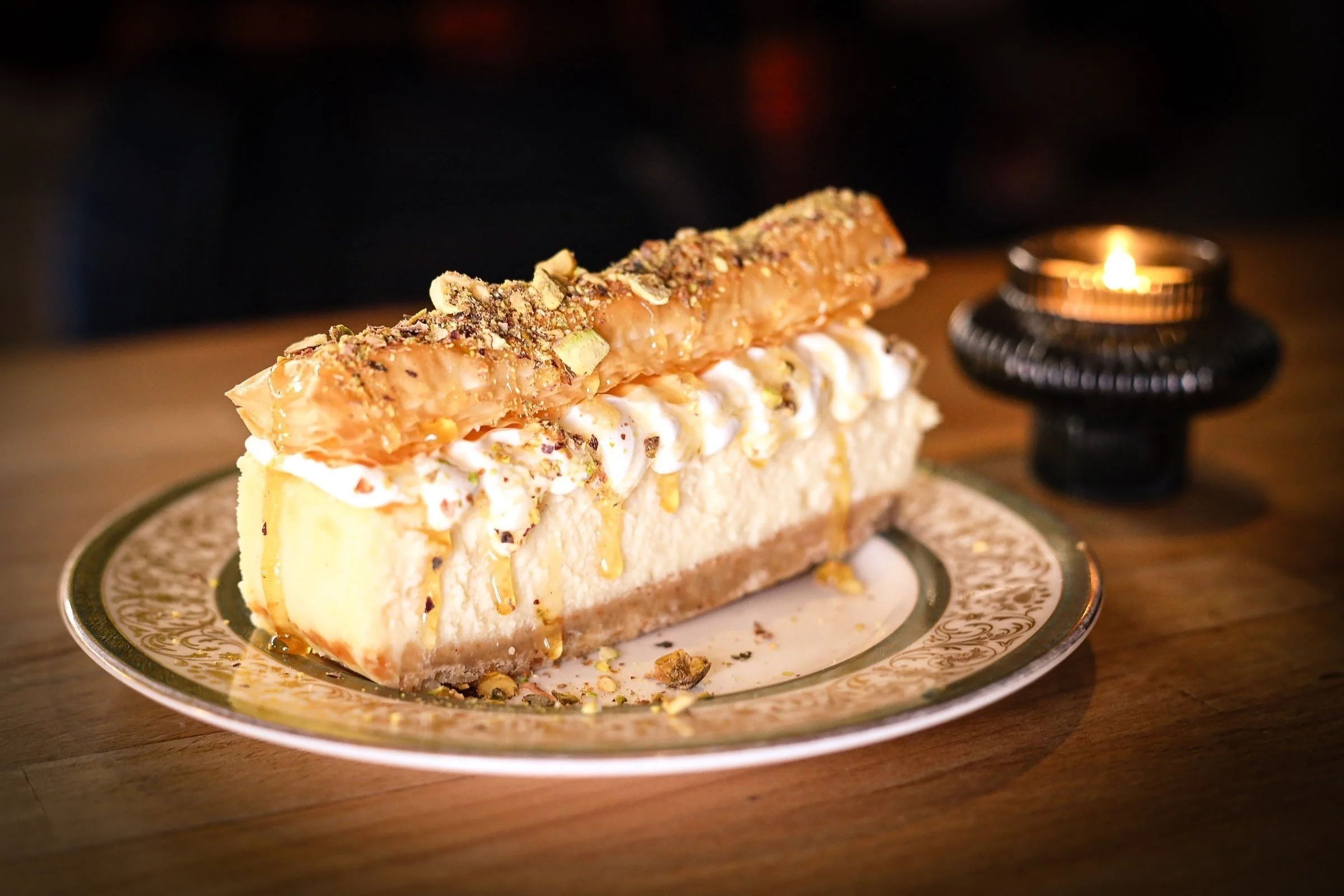 A slice of cheesecake topped with whipped cream, caramel sauce, chopped nuts, and a pastry stick is served on a decorative plate on a wooden table, with a lit candle in the background.