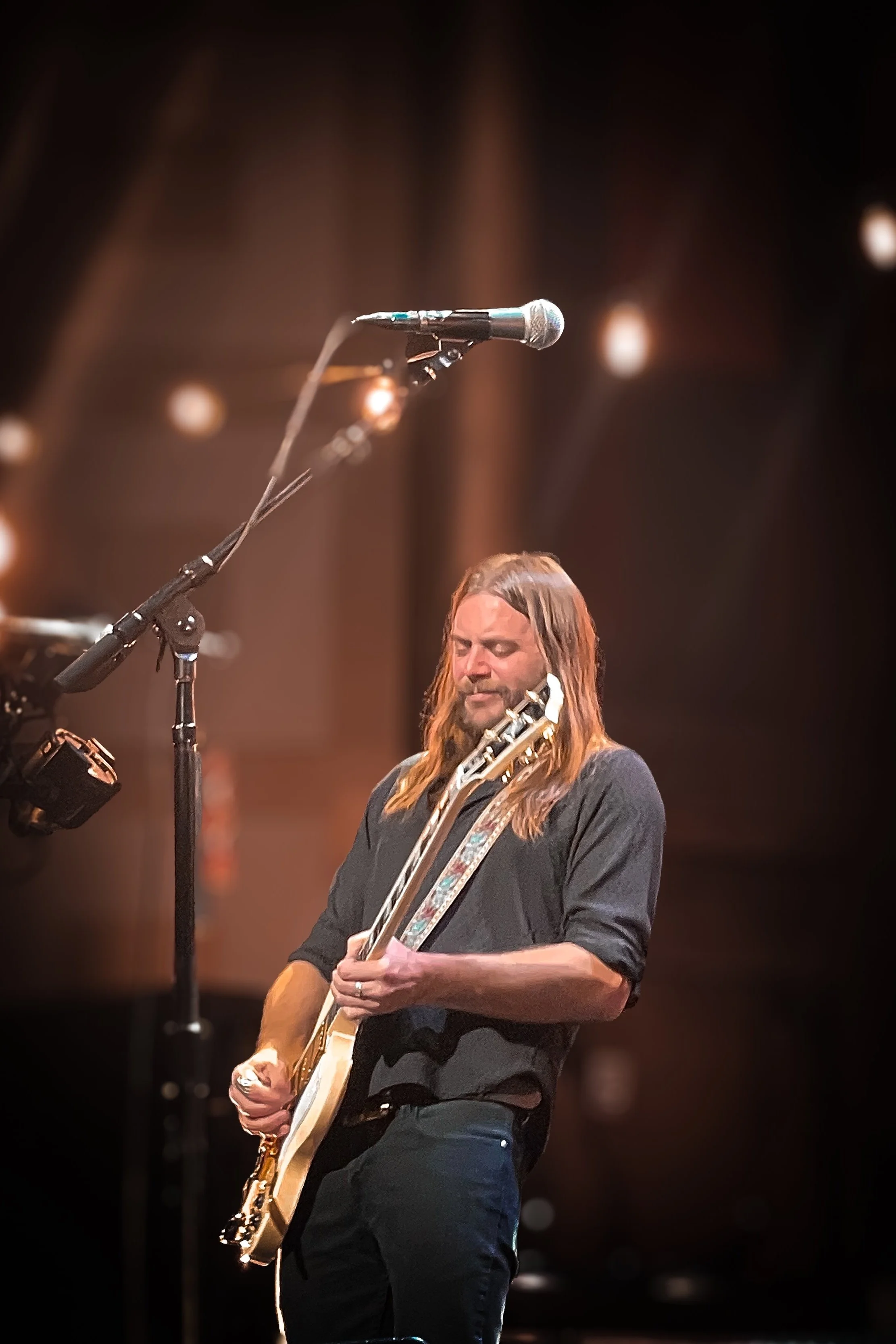 Male guitarist with long hair performing on stage with a microphone overhead and warm stage lighting.