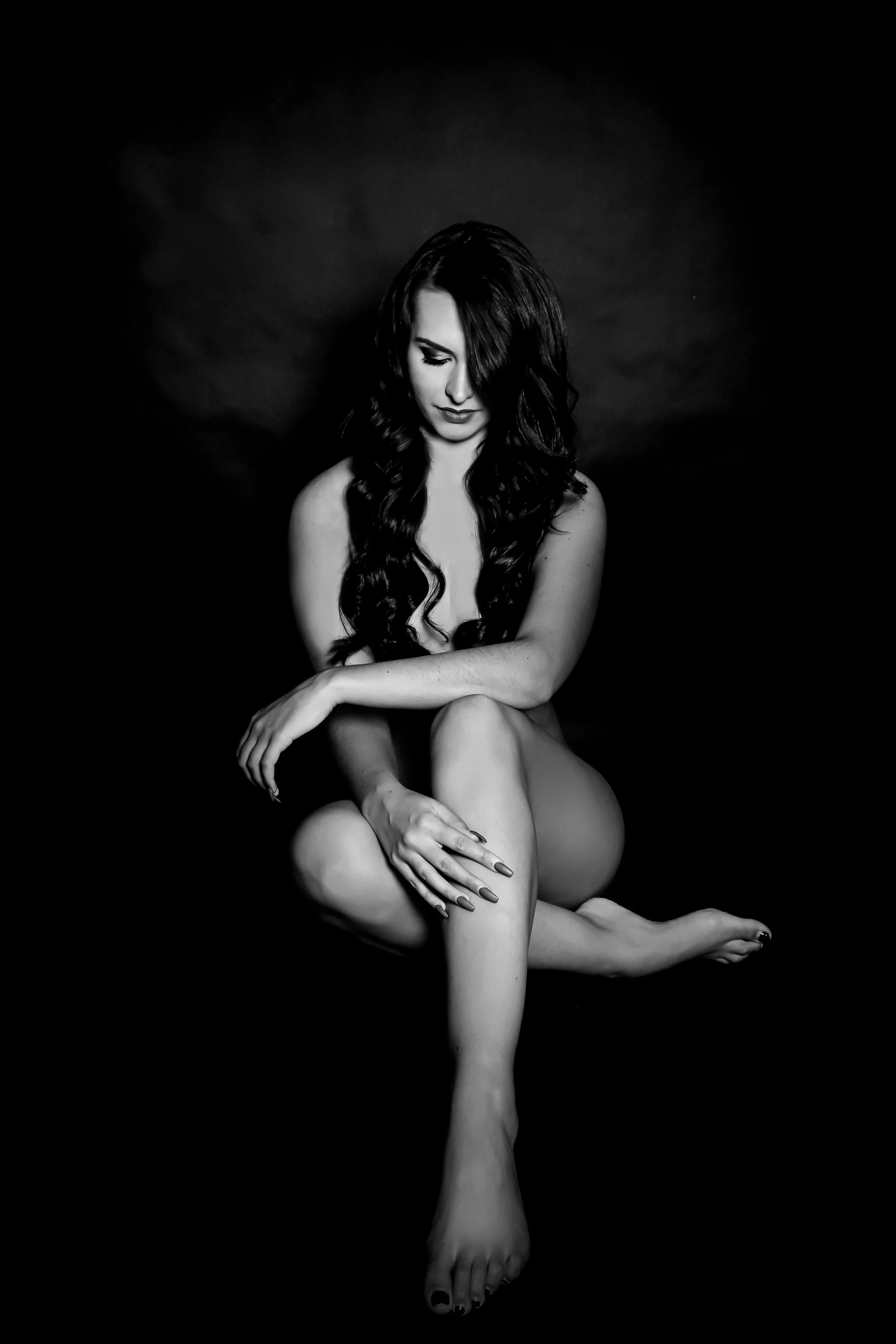 A black and white photograph of a woman sitting with her legs crossed, her head bowed, and eyes closed, against a dark background.