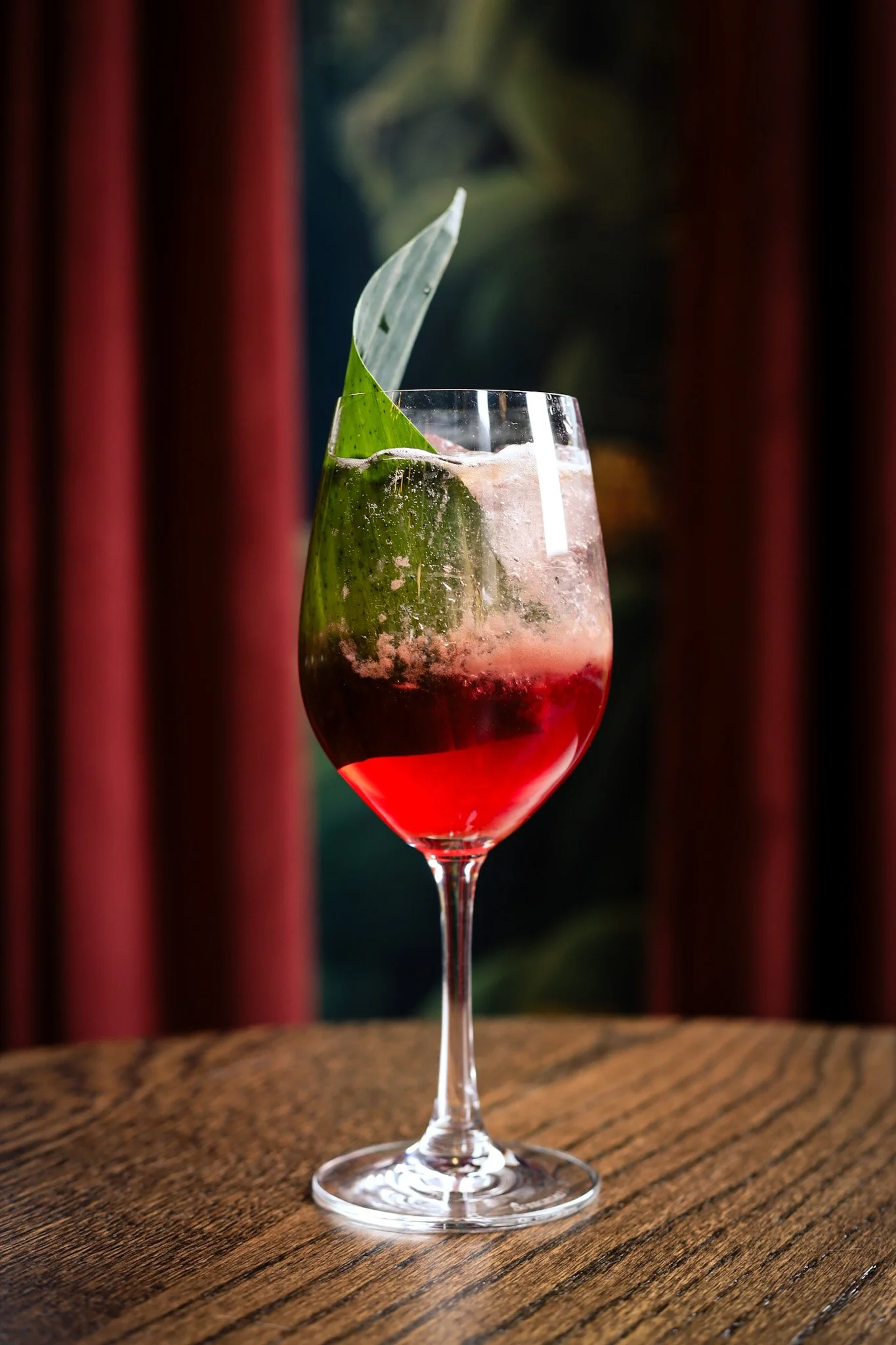 A tall wine glass with red and clear layered cocktail, garnished with a green leaf, on a wooden surface with a dark background and red curtains.