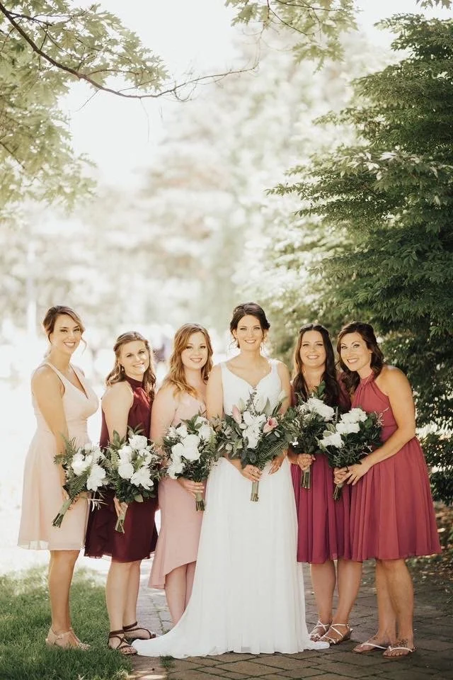 A bride in a white wedding dress standing with five bridesmaids in various shades of pink and maroon dresses, holding bouquets of white flowers, outdoors under trees.