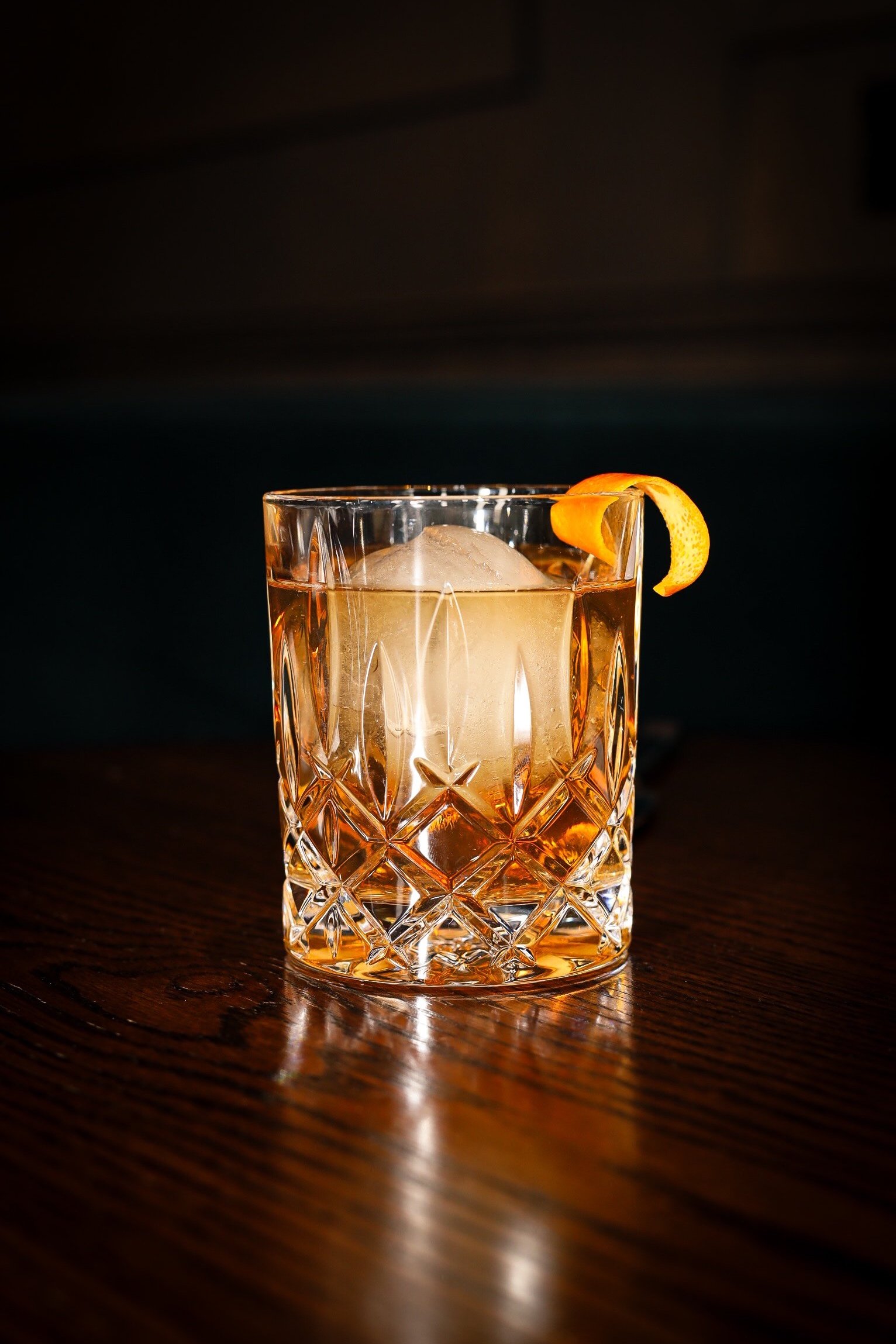 A glass of whiskey with a large ice cube and an orange peel garnish on a dark wooden surface.