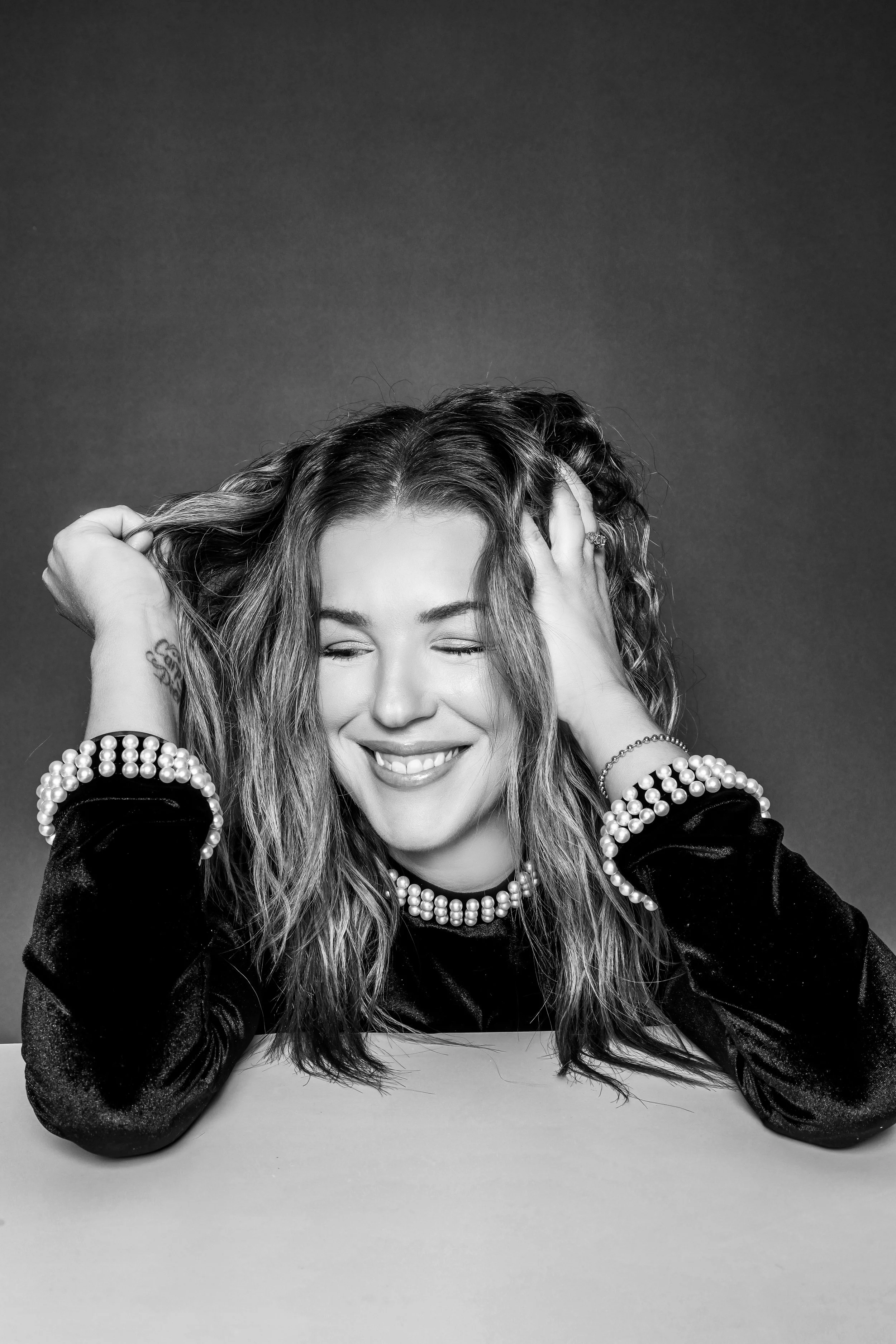 A woman with wavy hair smiling with eyes closed, wearing pearl jewelry, resting her elbows on a table, touching her head with one hand and holding hair with the other, in a black and white portrait.