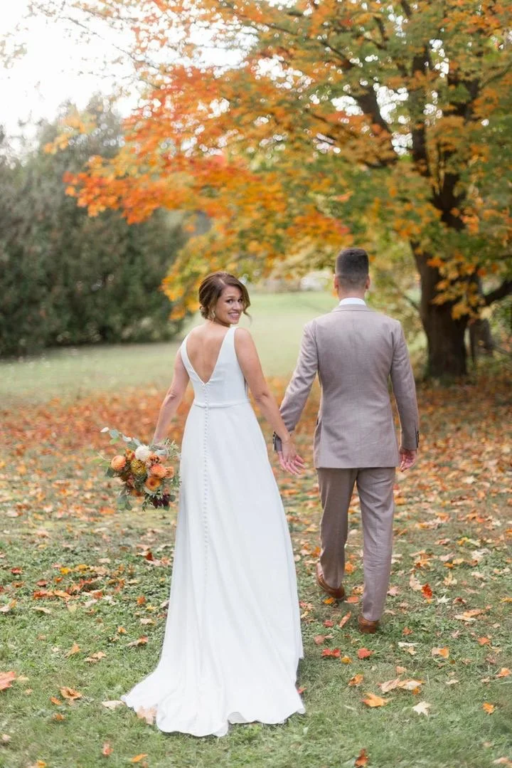 A bride and groom holding hands and walking in a park during autumn; the bride is in a white dress holding a bouquet, and the groom is in a gray suit with fall foliage in the background.