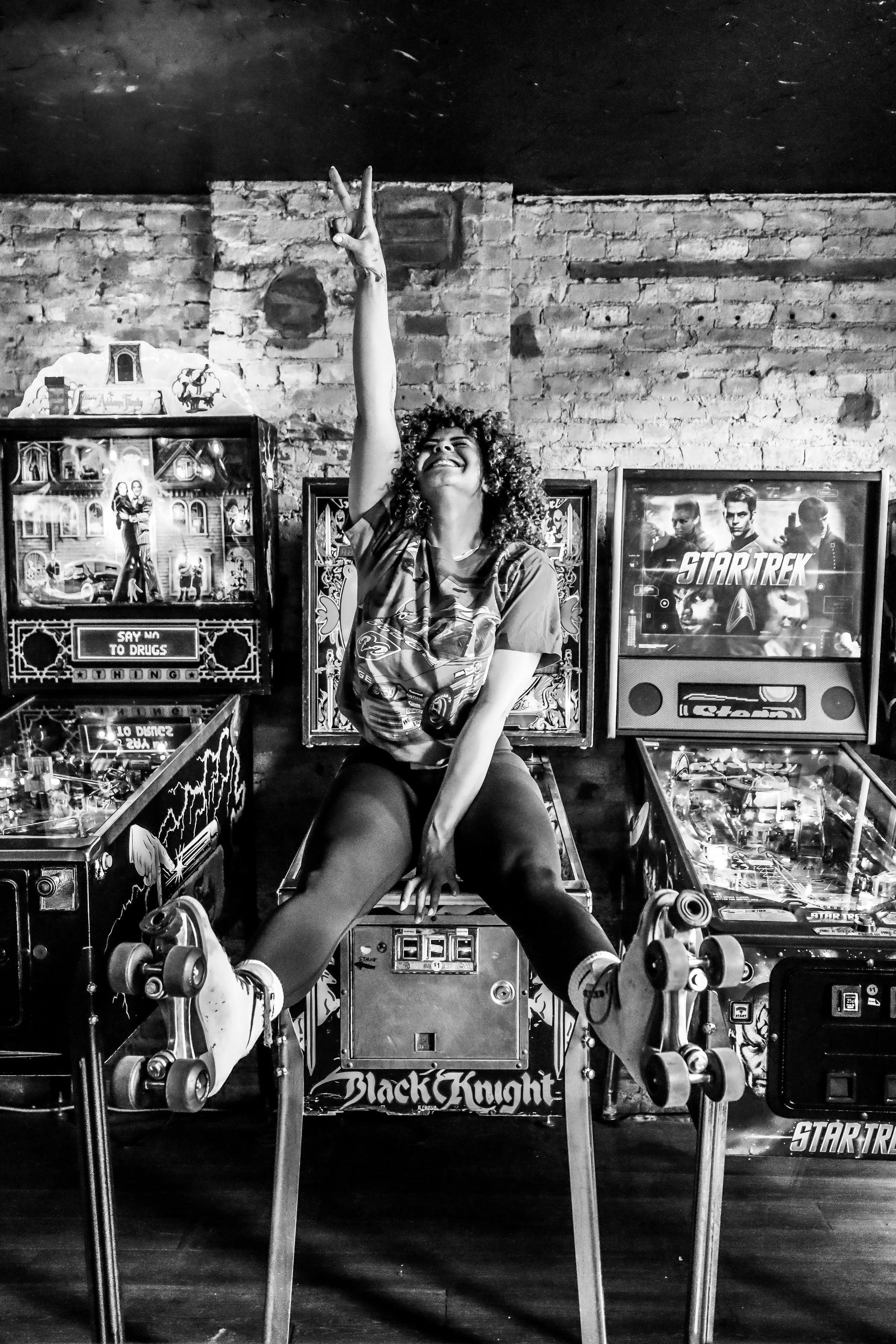 Woman with curly hair smiling and making a peace sign with her right hand while sitting on a pinball machine in a room with brick wall and framed artwork, including a Star Trek poster.