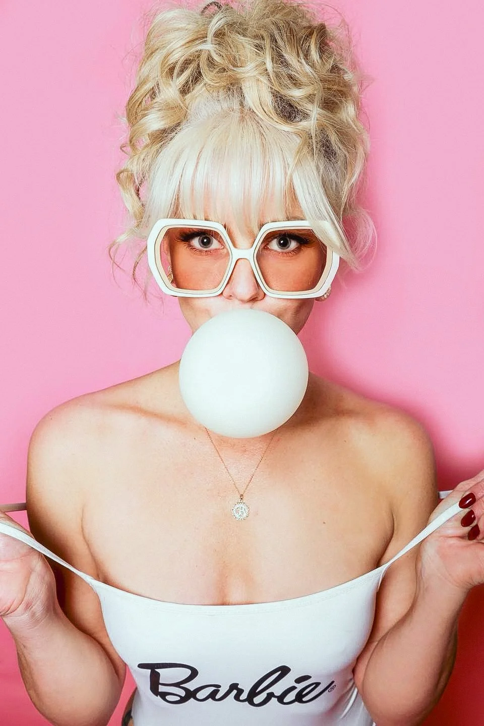 A woman with curly blonde hair and wearing large white sunglasses is blowing a bubble with bubblegum against a pink background. She is wearing a white off-shoulder top with the word 'Barbie' on it and a necklace with a pendant.
