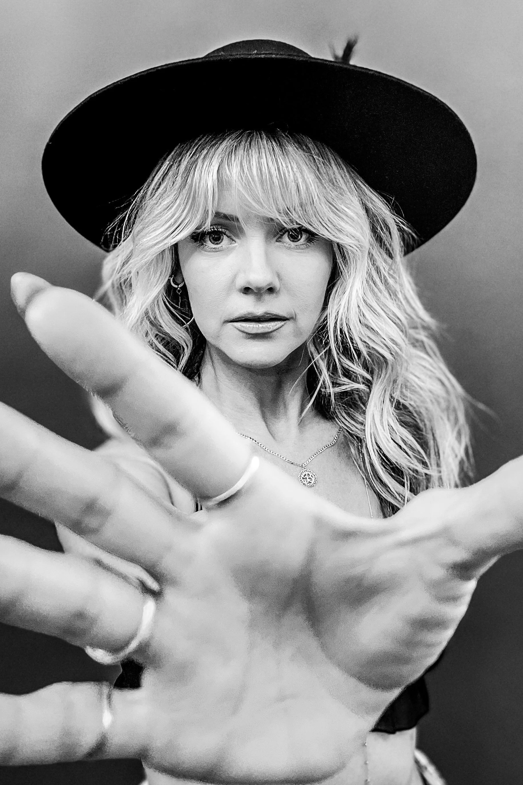 A Black and white photo of a woman with wavy hair wearing a wide-brimmed hat, looking directly at the camera with her hand reaching towards the lens.