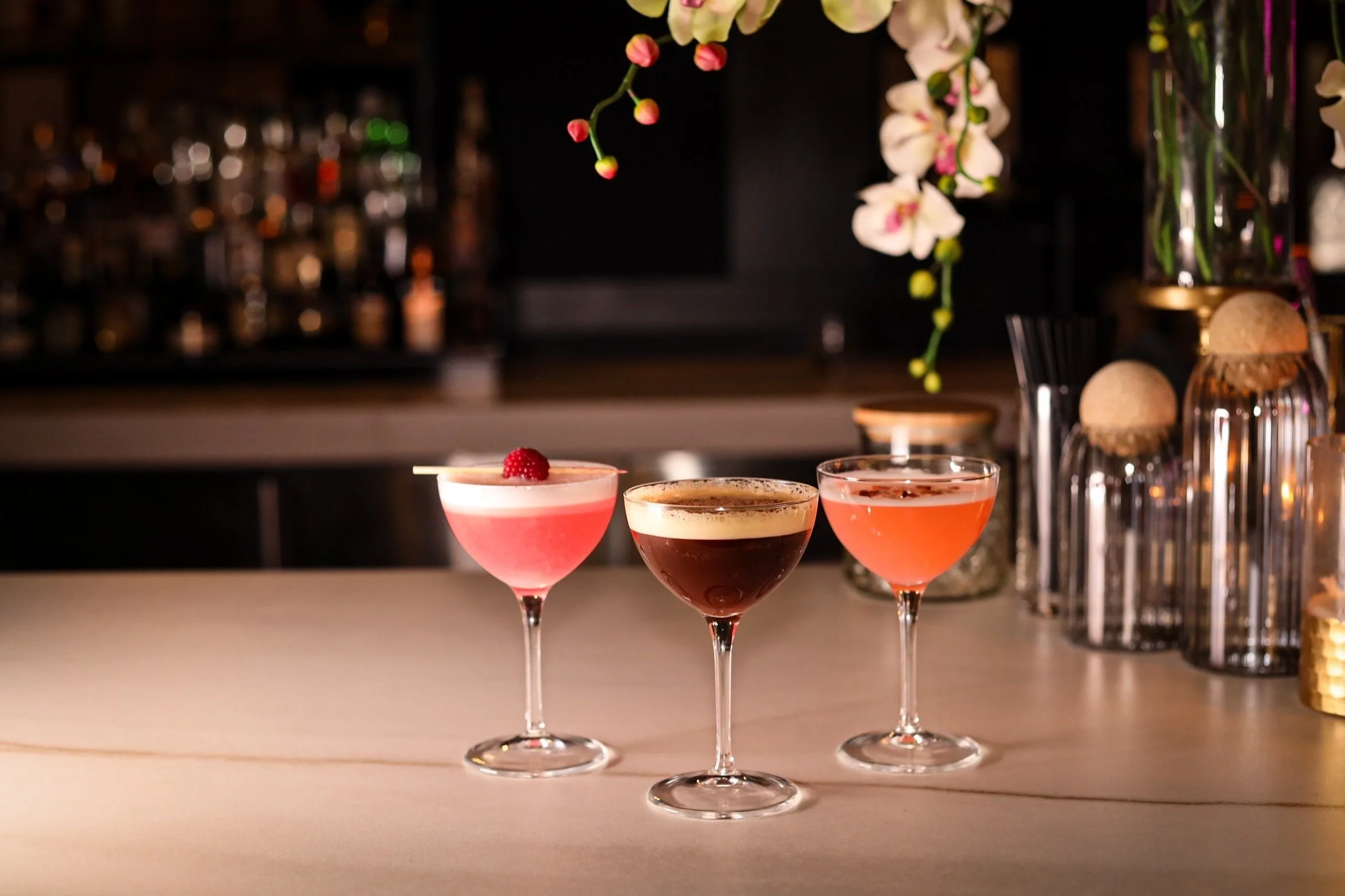 Three cocktails on a bar counter with a bar backdrop and decorative vases with flowers.