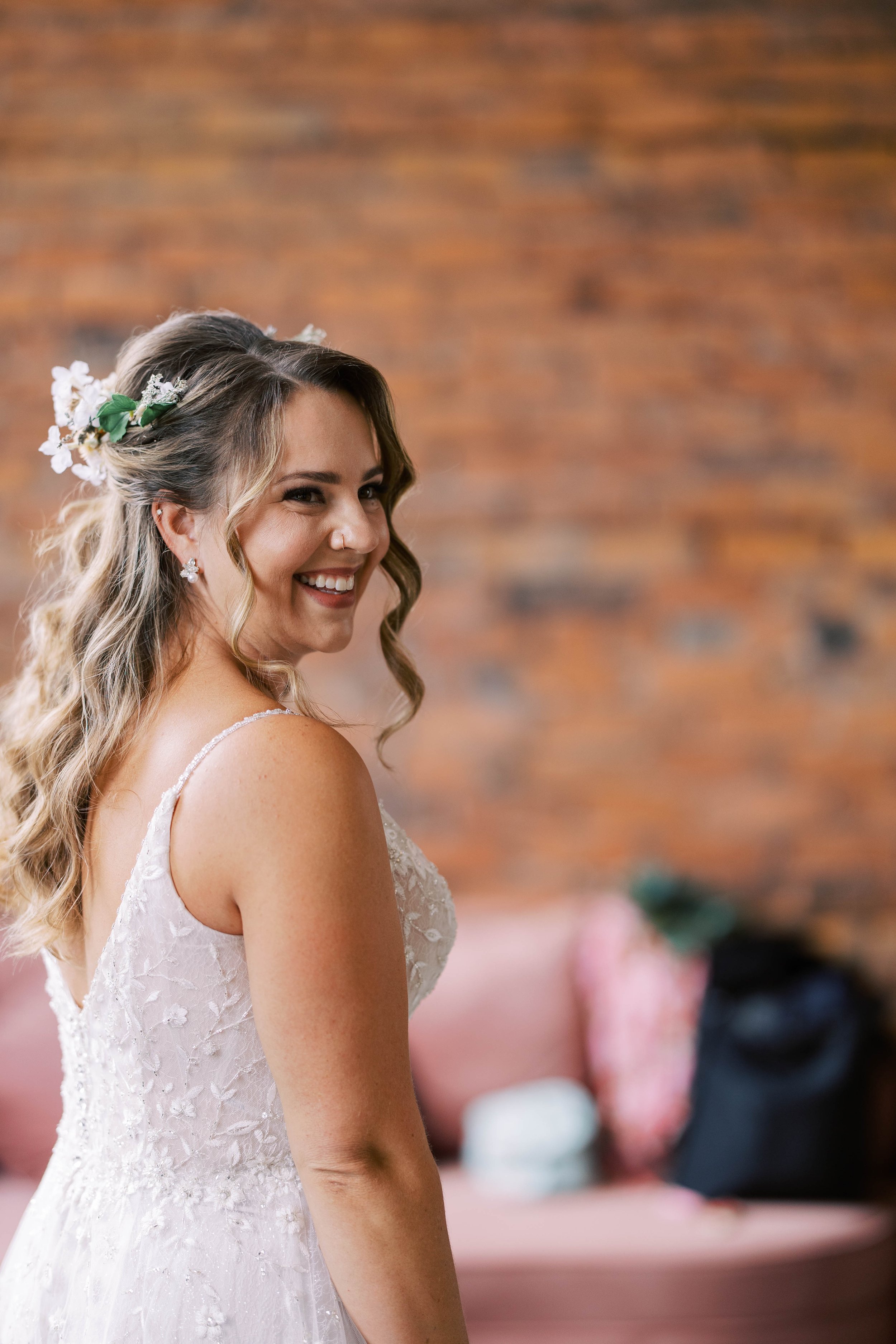 A smiling woman in a white wedding dress with floral hair accessories, standing indoors with a brick wall background.