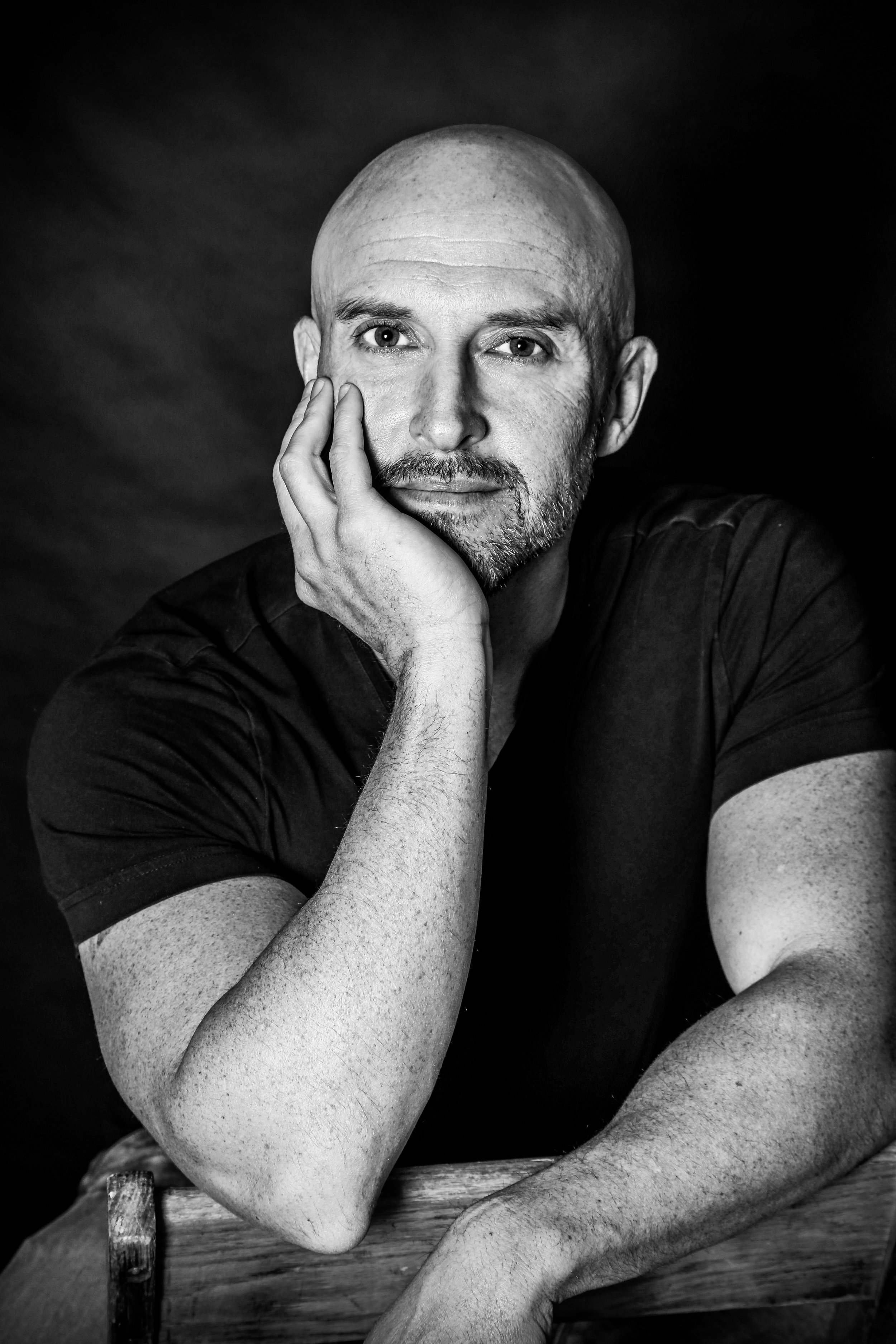 Black and white portrait of a bald man with a beard, resting his face on his hand, wearing a black T-shirt, looking directly at the camera.