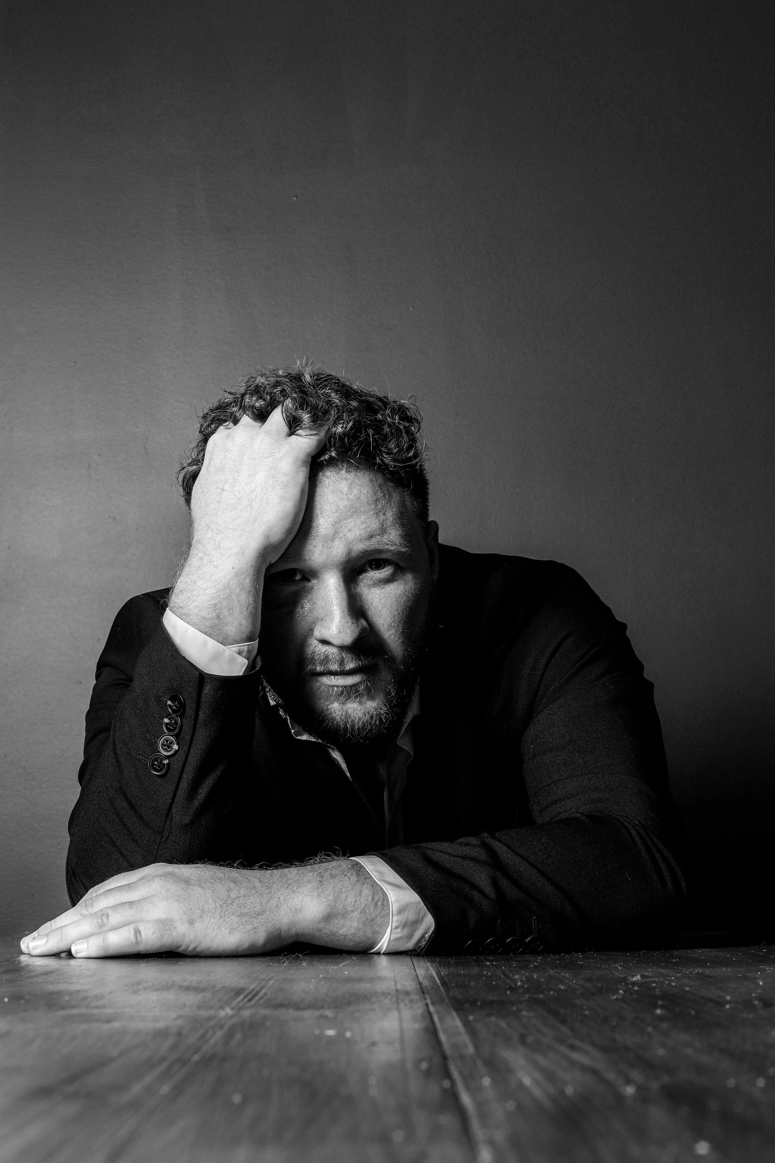 A black-and-white portrait of a man with curly hair and a beard, wearing a dark suit, resting his head on his left hand, leaning on a wooden table, with a plain background.