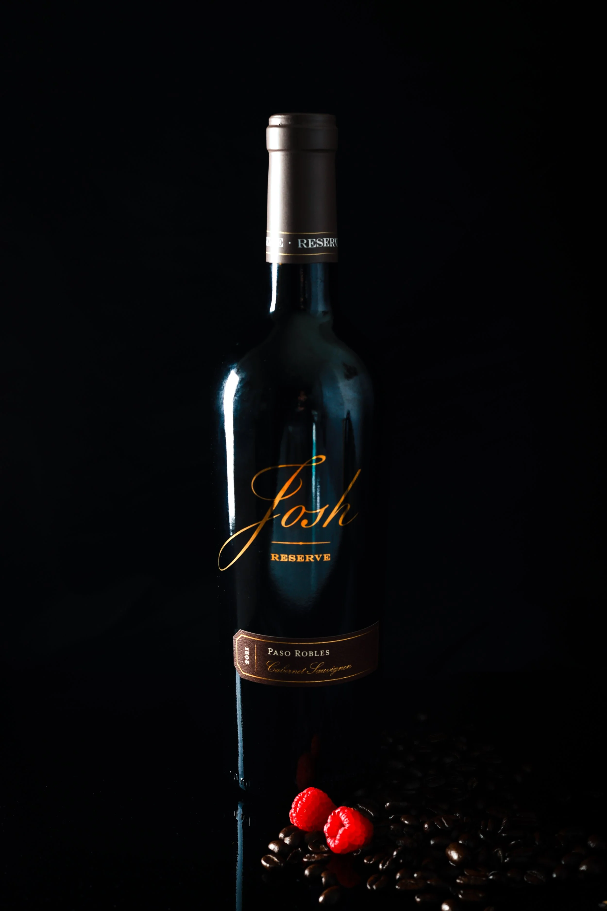 A dark-colored wine bottle labeled "Josh Reserve" with a black background, positioned next to scattered coffee beans and three red raspberries at the bottom of the image.