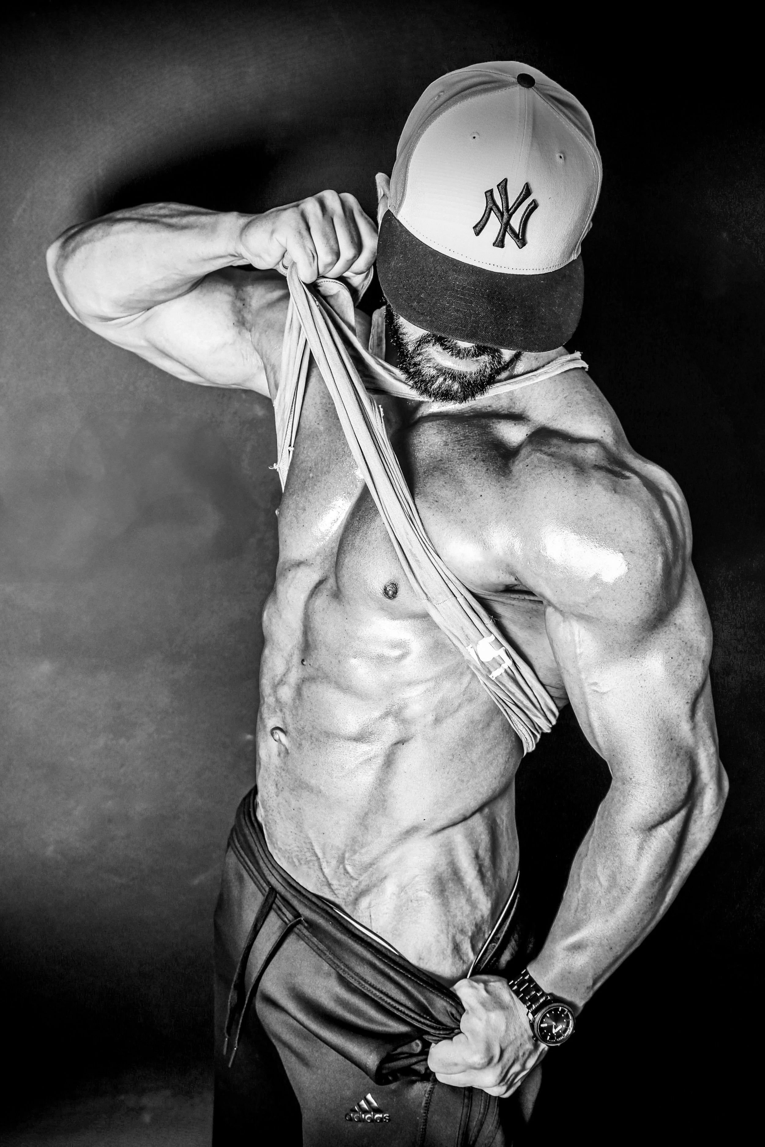 Black and white photo of a muscular man lifting his shirt to show his abs, wearing a New York Yankees cap and athletic pants, against a dark background.