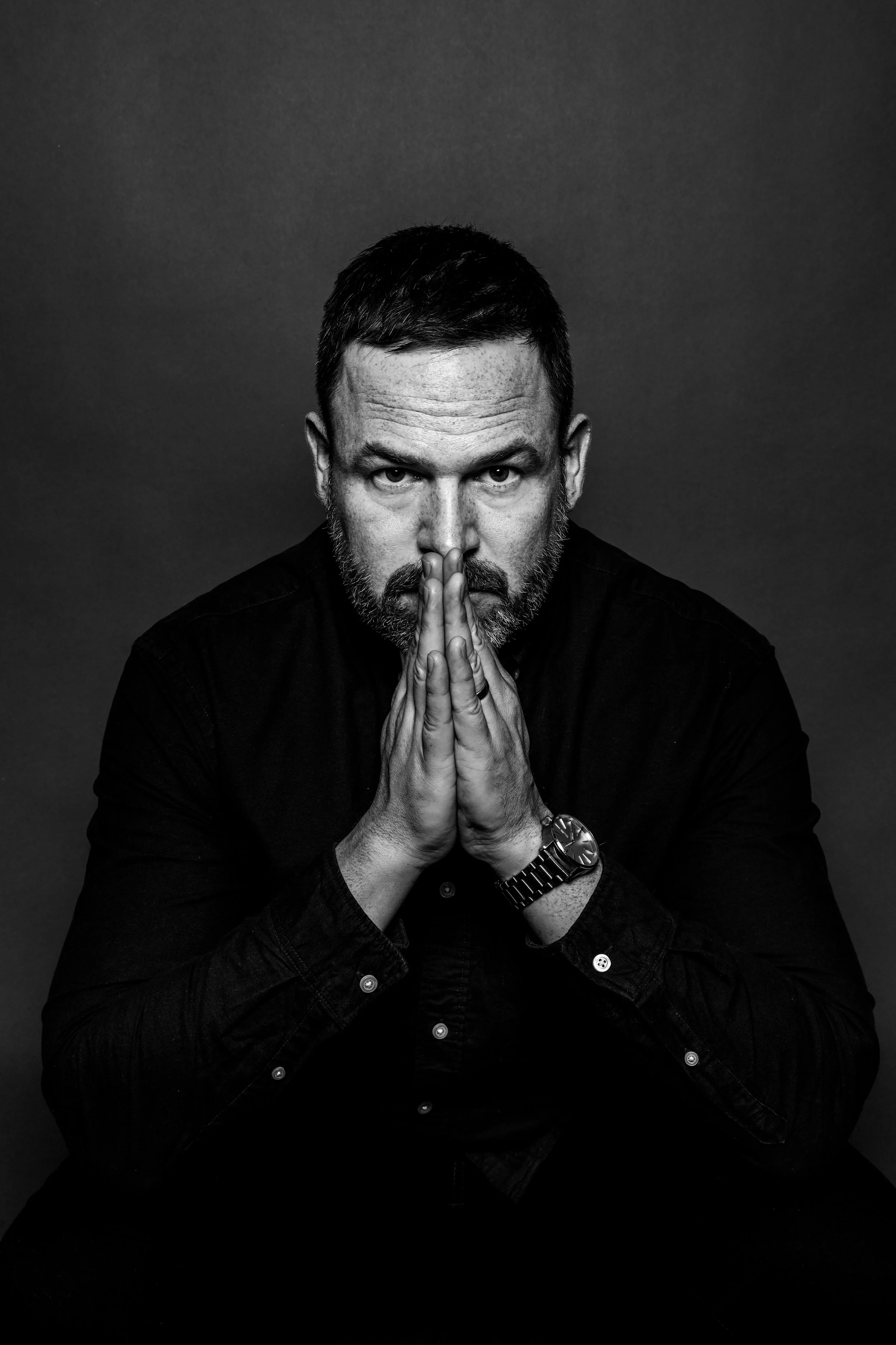 Black and white portrait of a man with short dark hair and a beard, wearing a dark shirt and a wristwatch, with his hands pressed together in front of his face, looking intensely at the camera against a plain dark background.