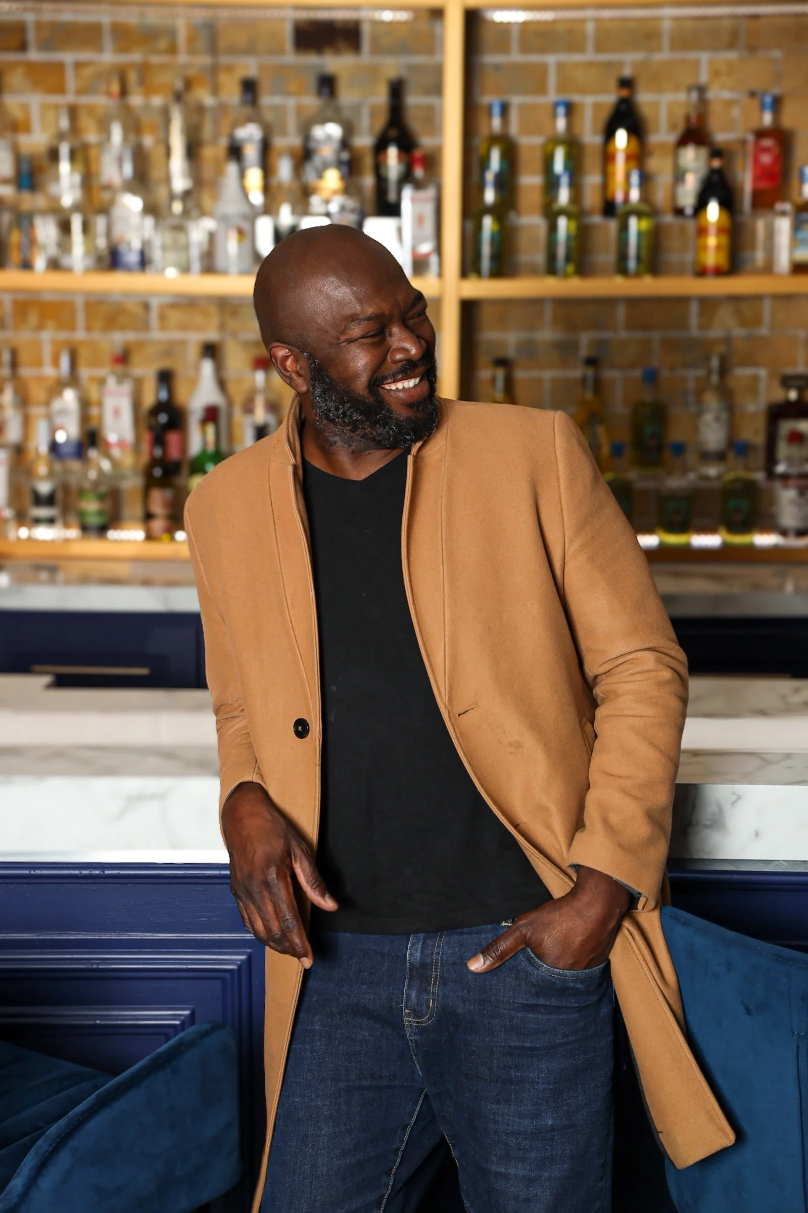 A man with a bald head and beard smiling and leaning against a bar in a lounge or bar area, with shelves of liquor bottles in the background.