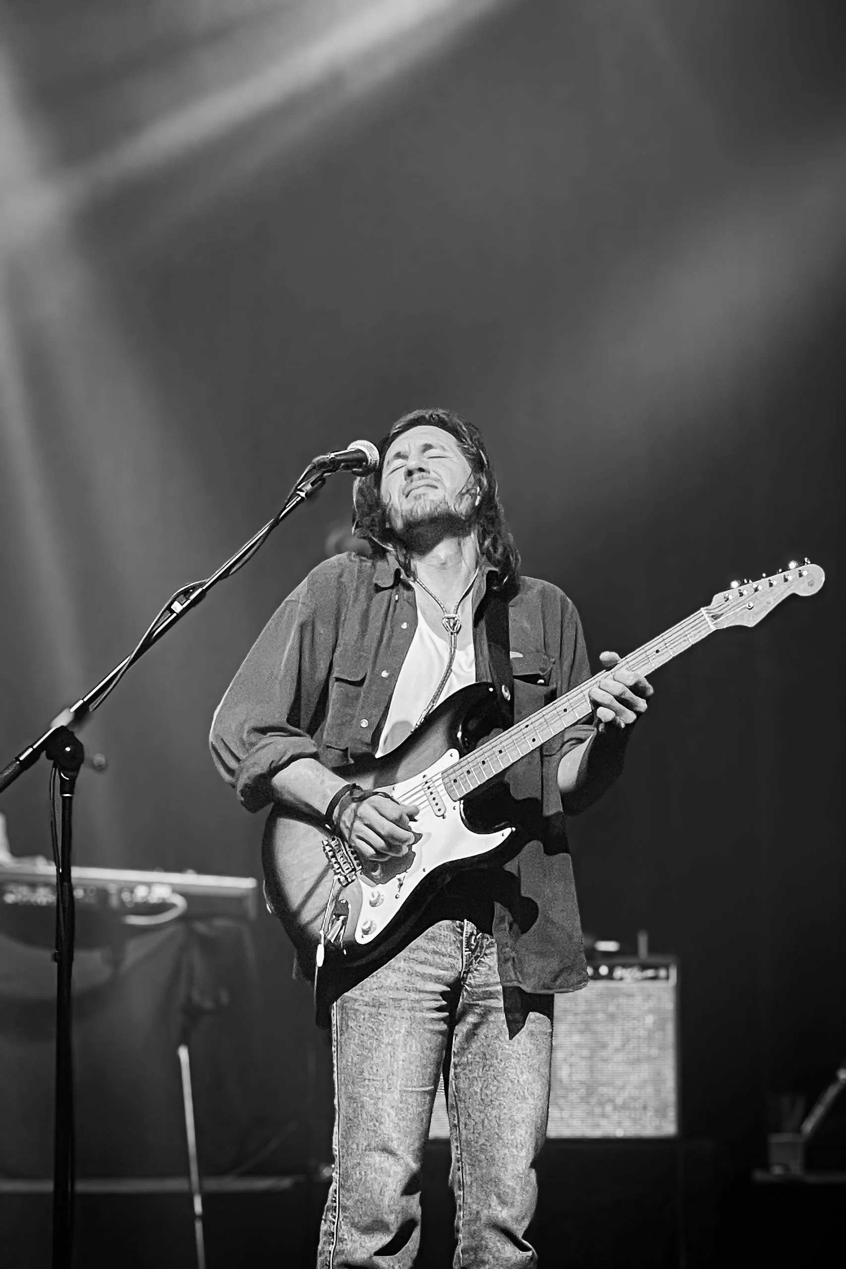 A man with long hair and a beard playing an electric guitar on stage with eyes closed and a joyful expression, black and white photo.