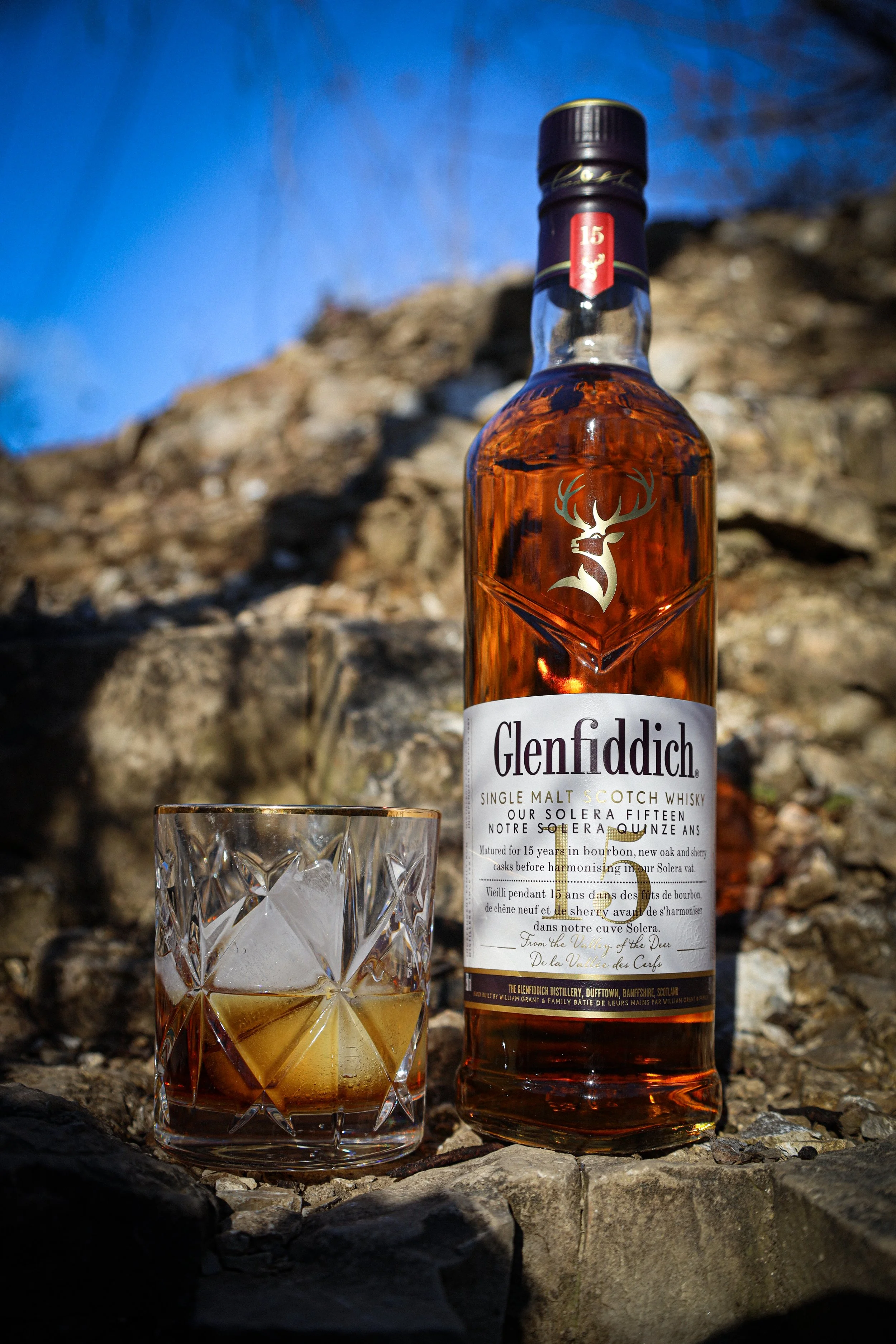A bottle of Glenfiddich single malt scotch whisky and a glass filled with whisky and ice, set outdoors on rocks with a blue sky background.