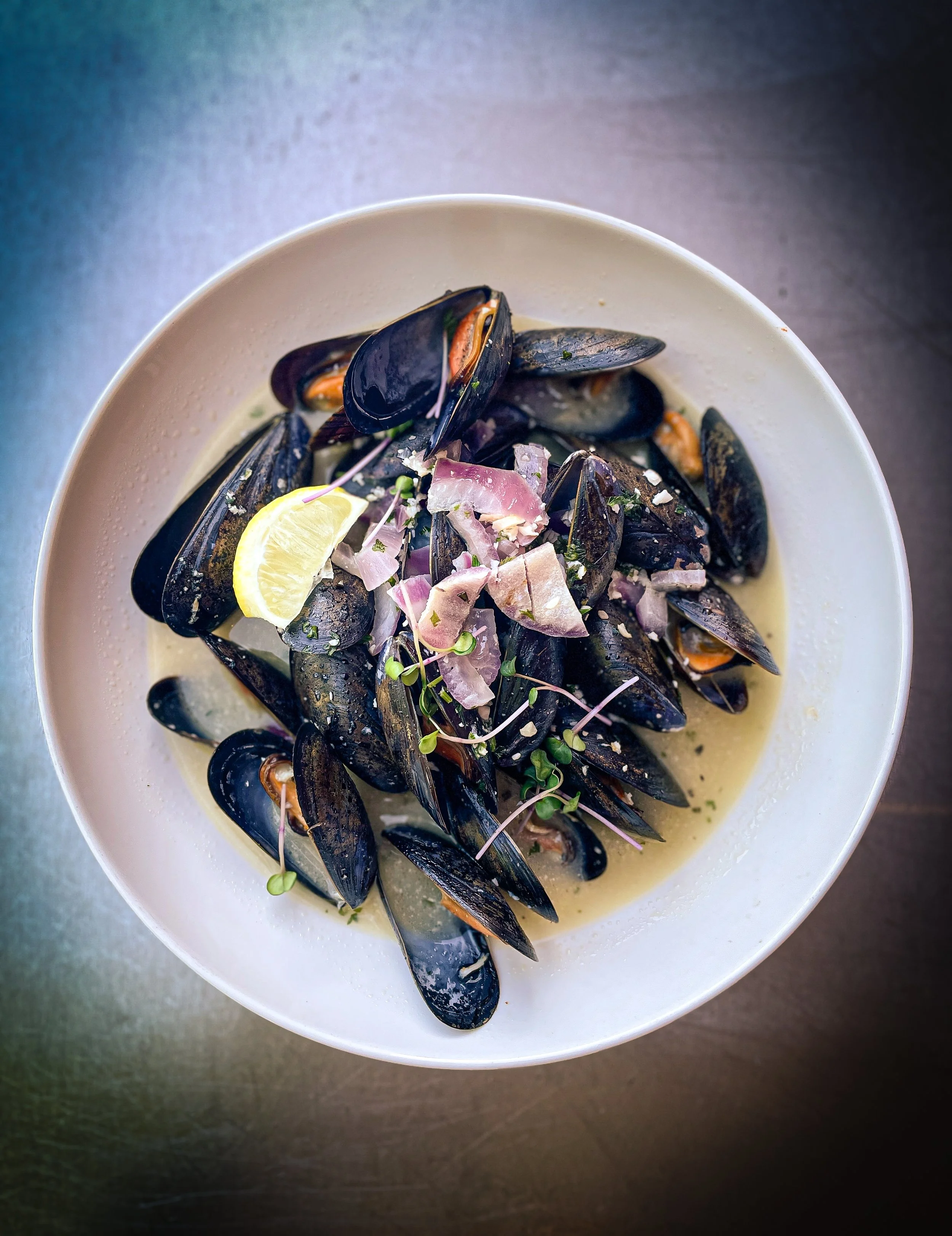 A bowl of steamed mussels in a creamy sauce, garnished with chopped onions, microgreens, and a lemon wedge.