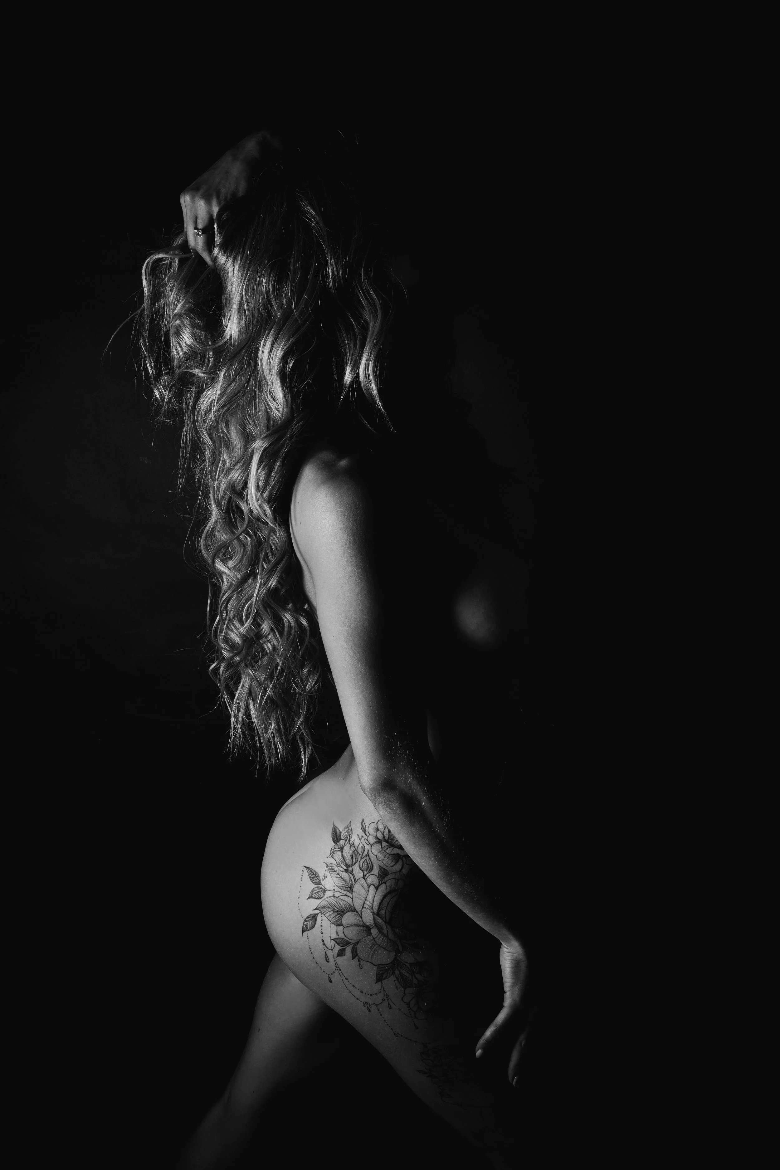 A black and white photo of a woman with long, wavy hair, covering her face with her hand, and a floral tattoo on her thigh.