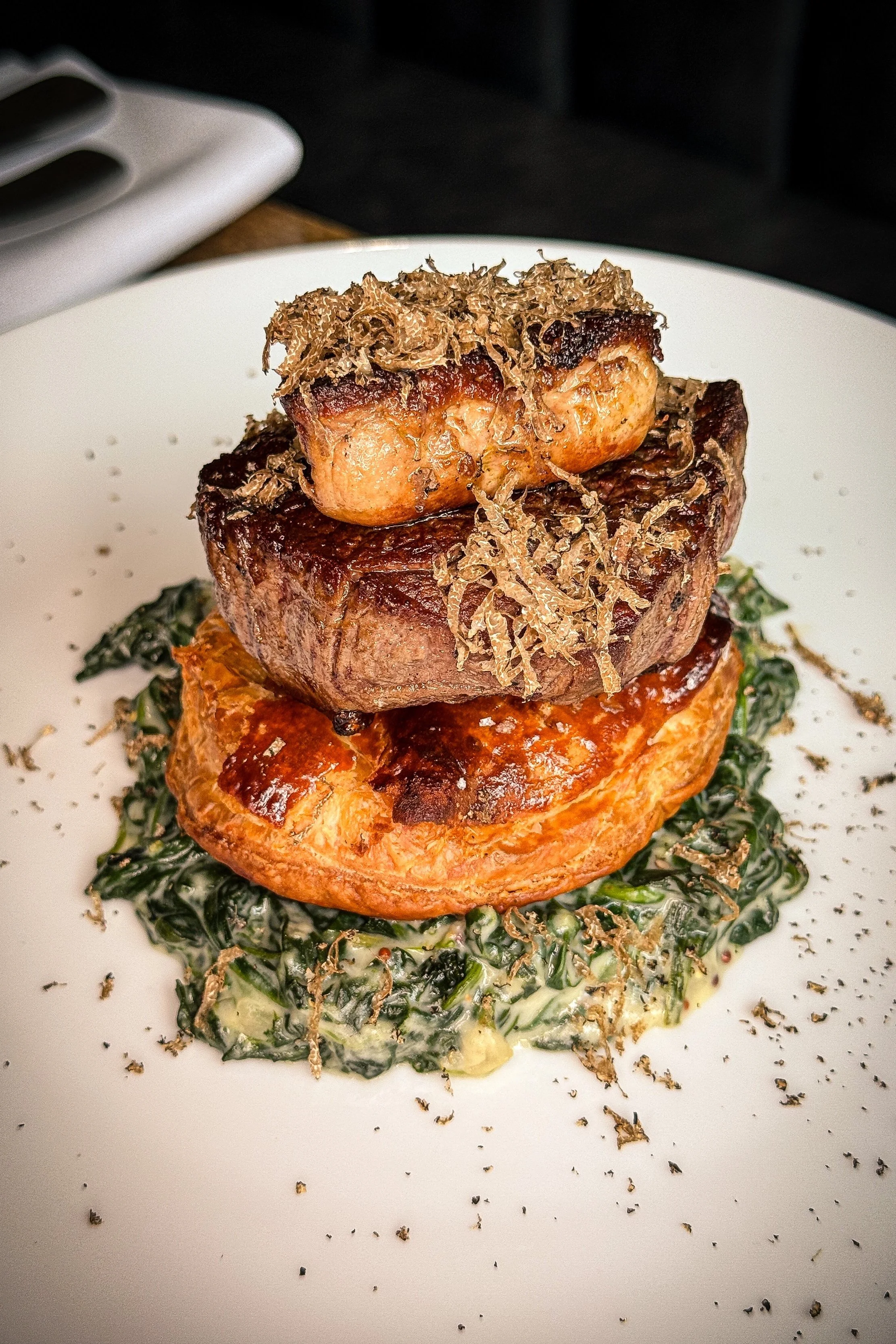 A plated gourmet beef dish with a seared steak topped with black truffle shavings and a poached egg, served over creamed spinach.
