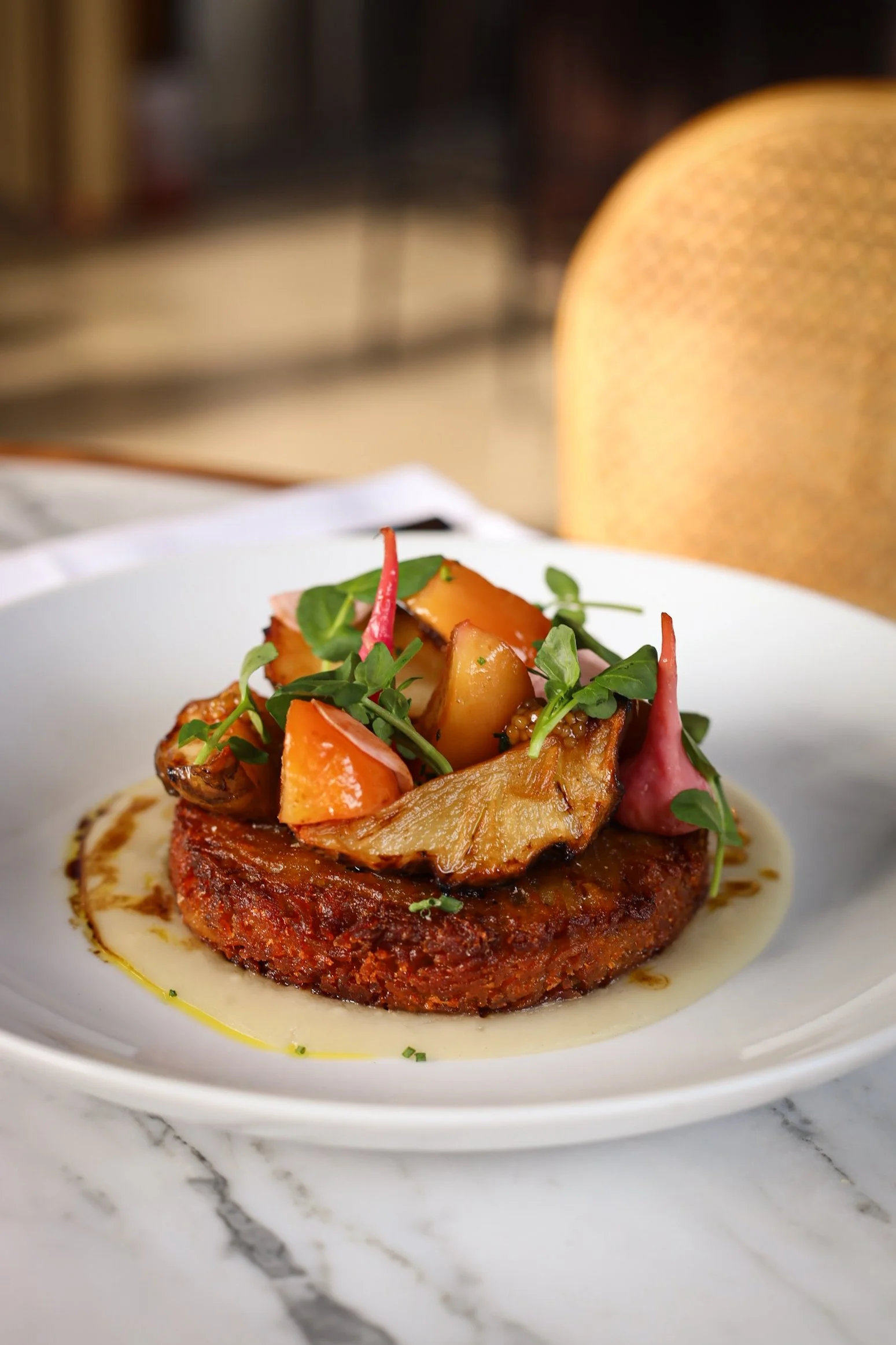 A gourmet dish featuring a hamburger patty topped with roasted vegetables and microgreens, served on a white plate with a creamy sauce on a marble surface.