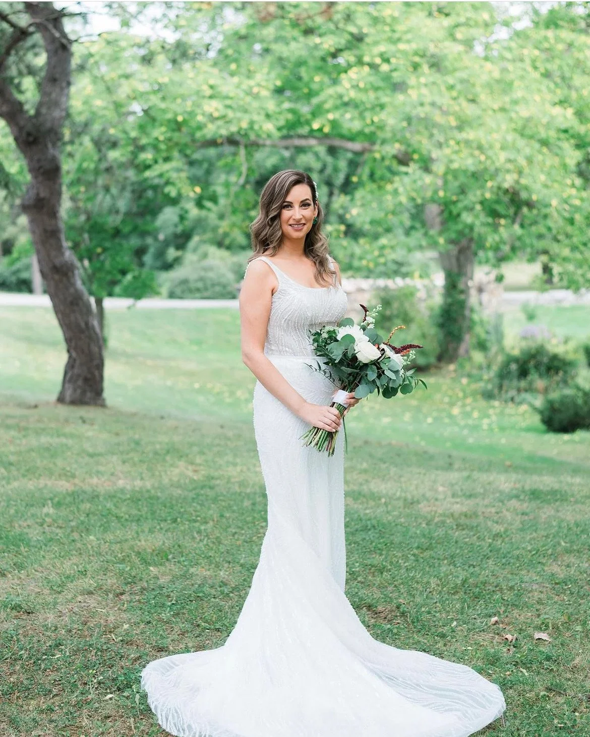 A woman in a white wedding dress standing on grass holding a bouquet of flowers, outdoors with green trees in the background.