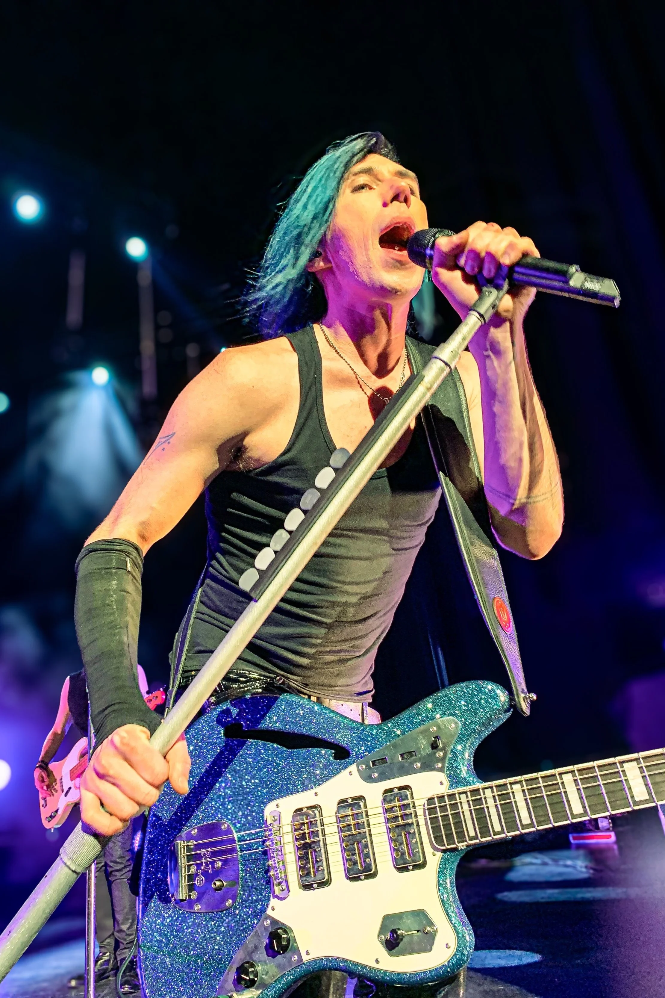 A musician with blue hair singing into a microphone on stage, wearing a black tank top and playing a blue glittery electric guitar.