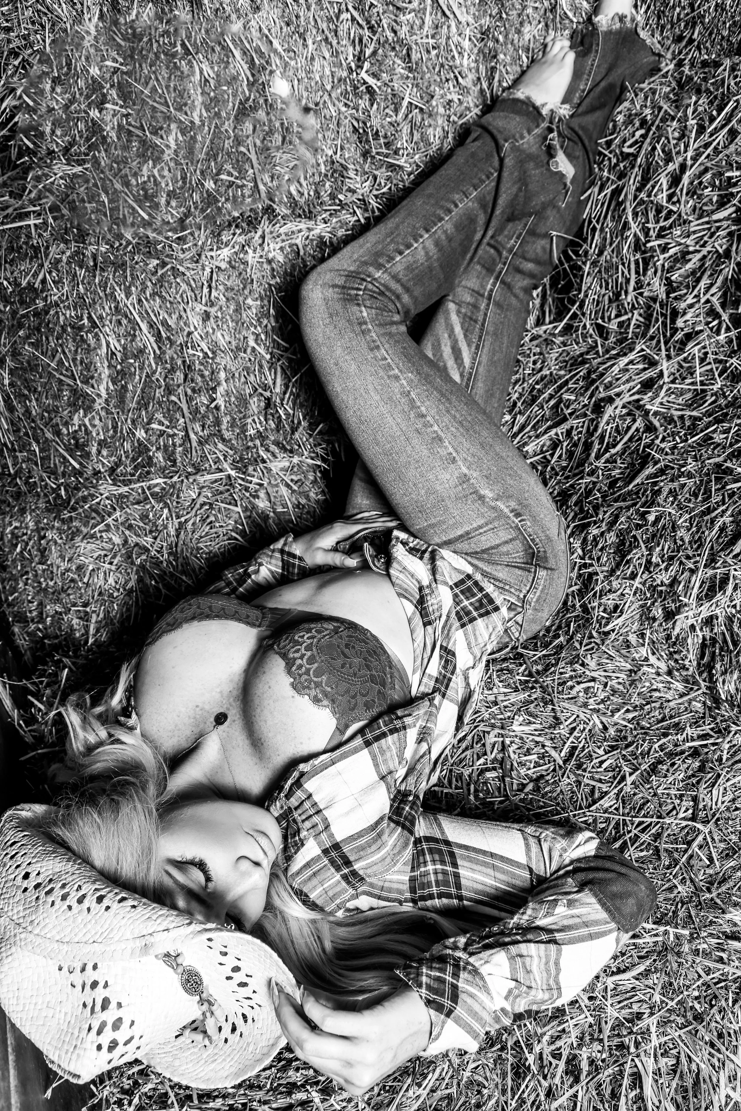 A woman lying on the grass, wearing jeans, a plaid shirt, and a lace bra, with a straw cowboy hat covering her face, in black and white photography.
