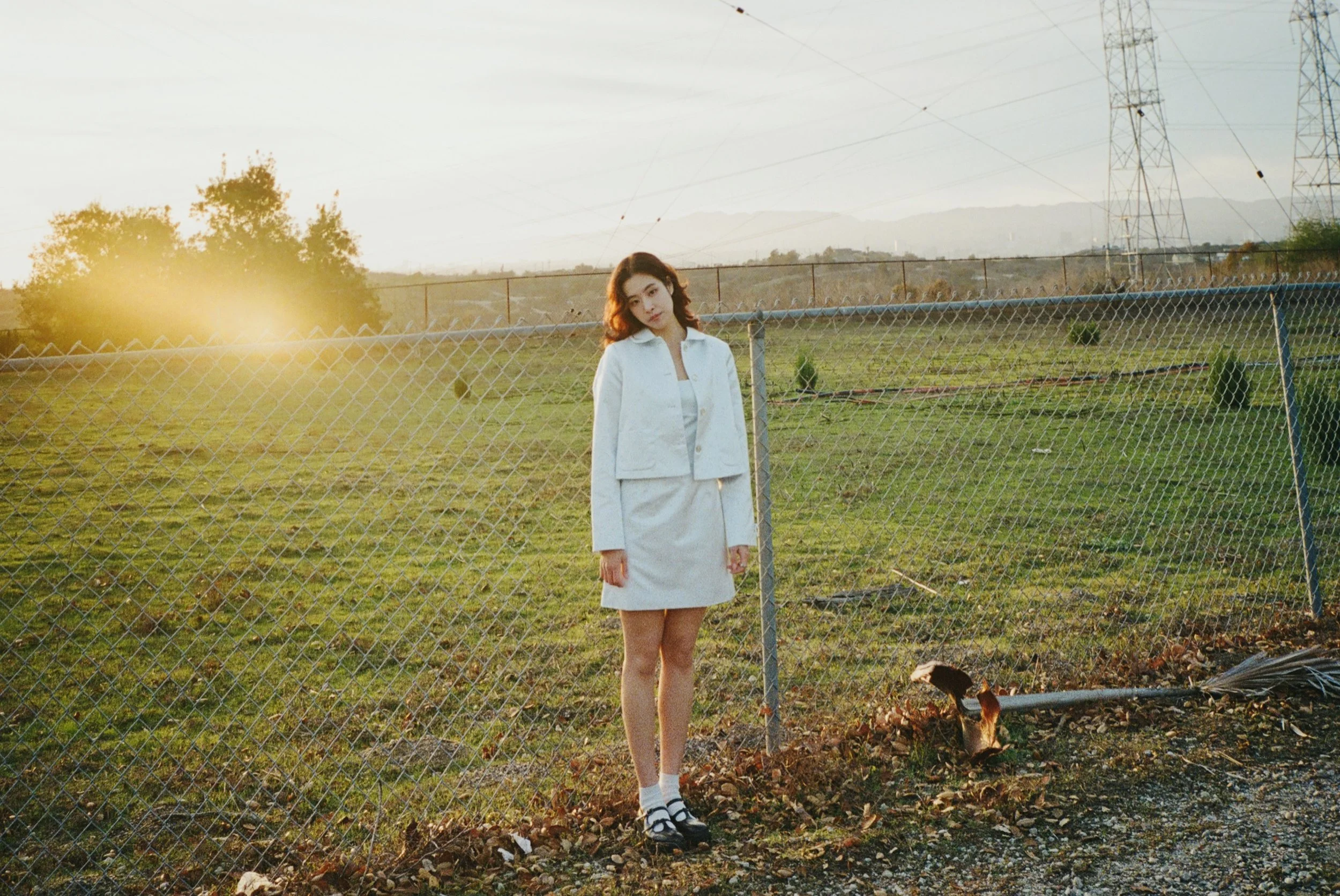 A young woman in a white outfit standing by a chain-link fence in an open field during sunset.