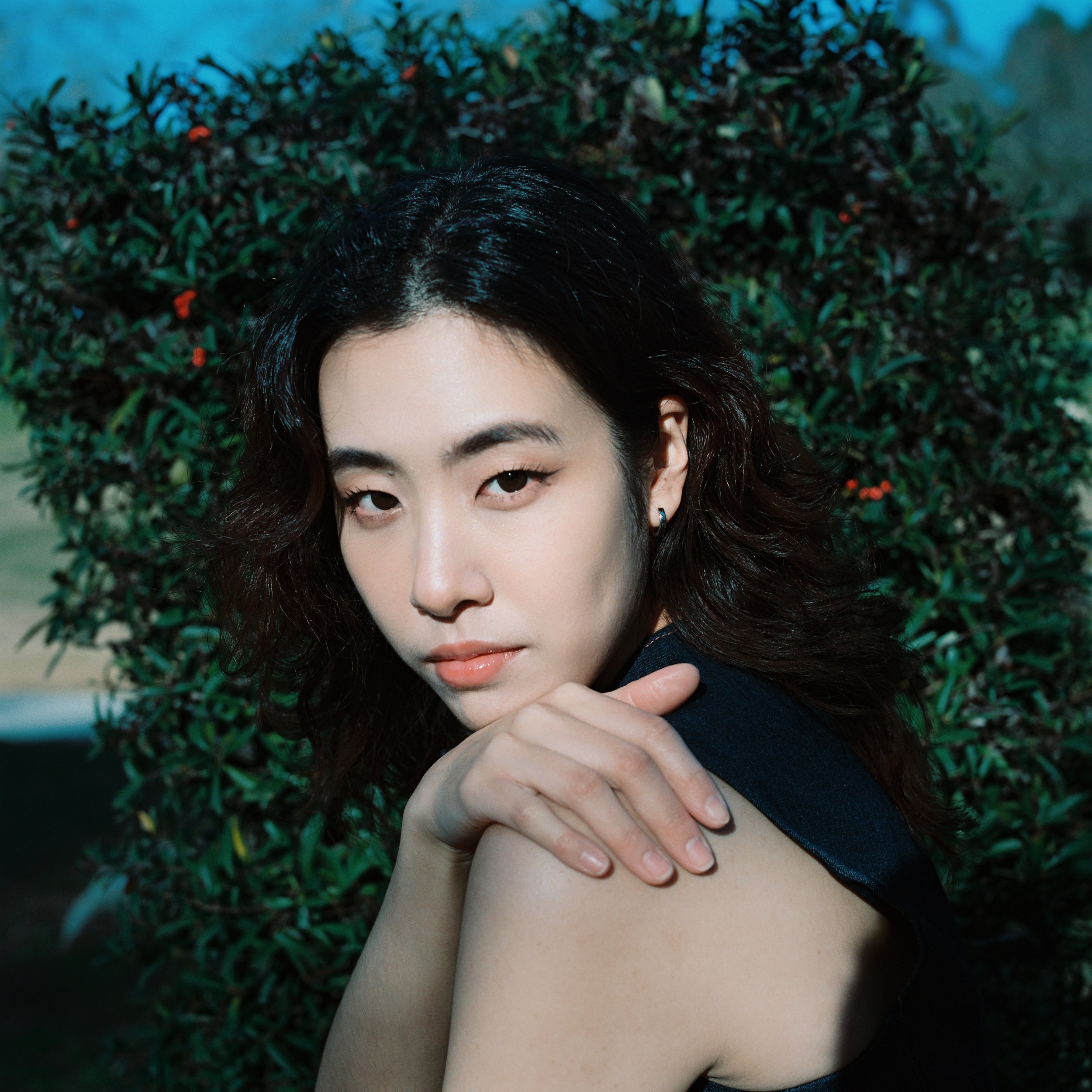 A woman with black wavy hair resting her head on her hand, looking at the camera with a confident expression, in front of green bushes.