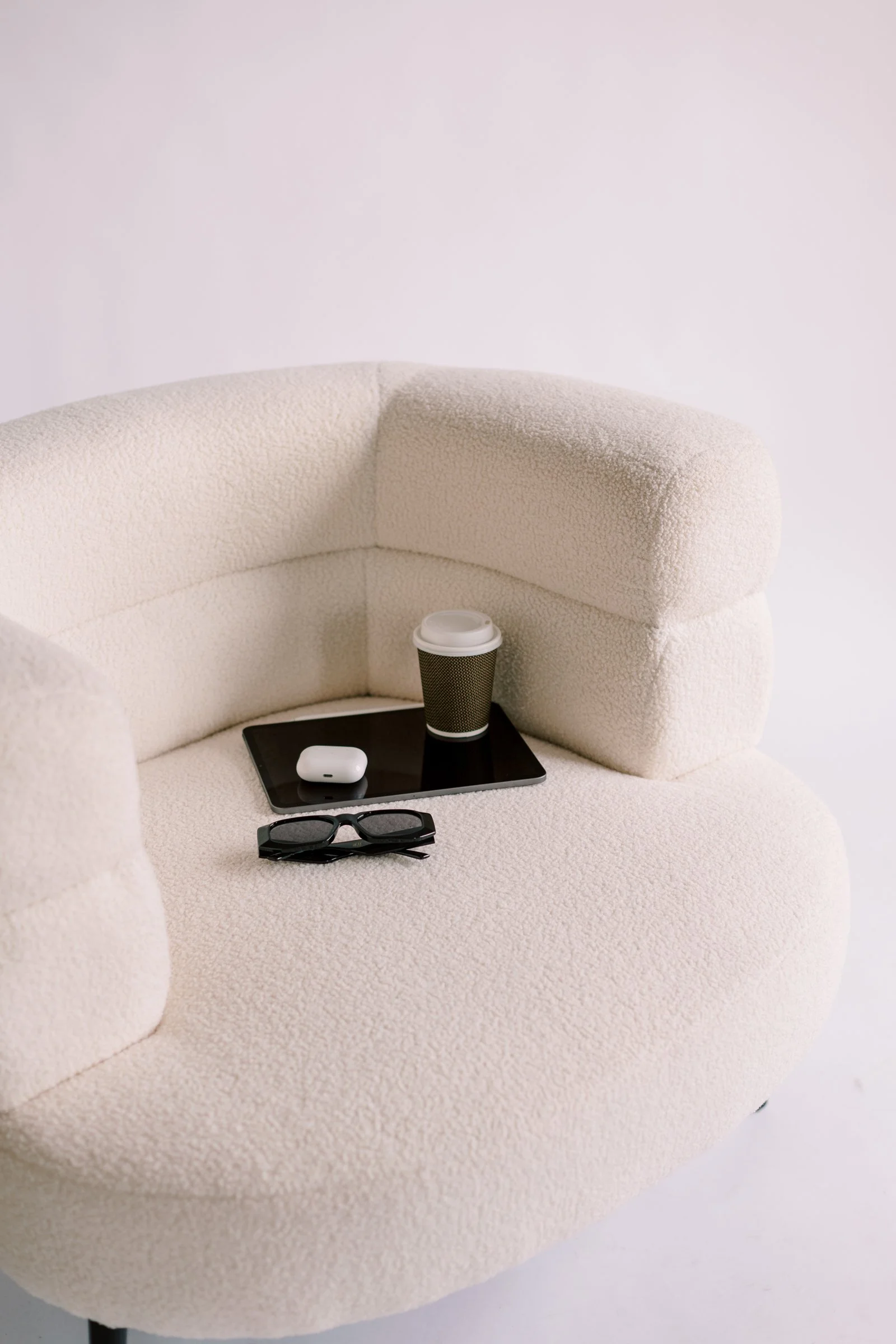 Rise2Freedom |  A cozy beige armchair with textured fabric, holding a pair of sunglasses, a tablet, a white wireless earbud case, a coffee cup with a black sleeve, and a black coffee lid.