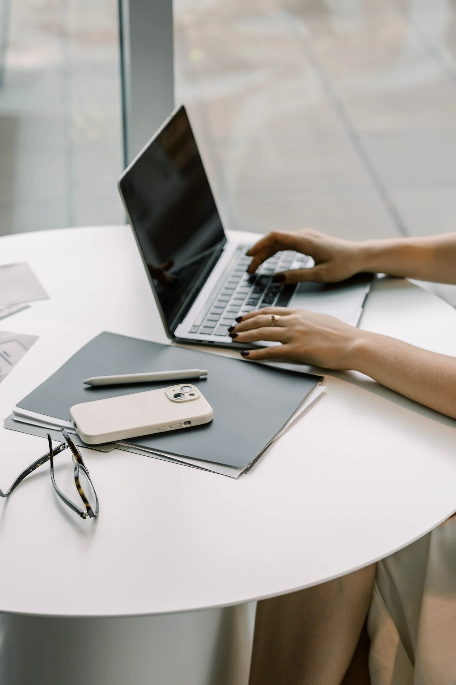 A person working on a laptop at a white round table with a smartphone, glasses, a pen, and notebooks.