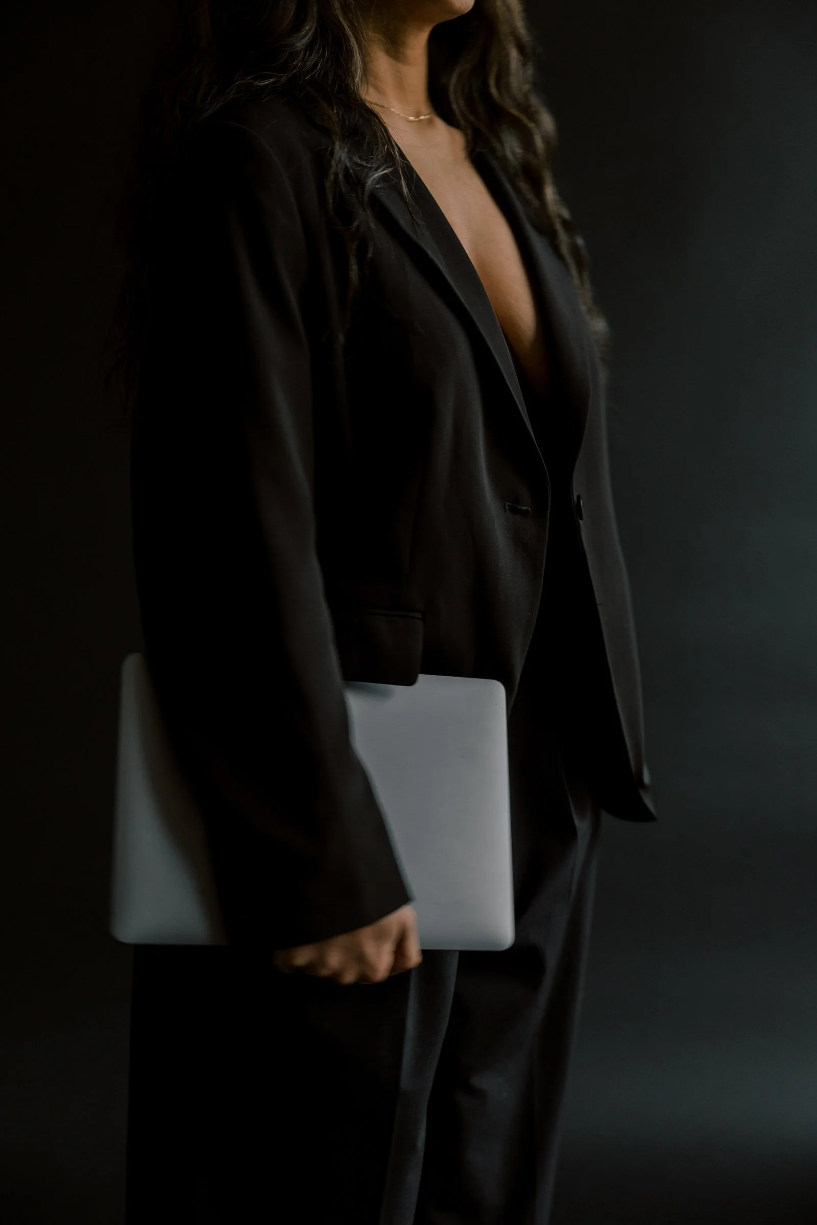 Woman in black suit holding a silver laptop, standing against a dark background.